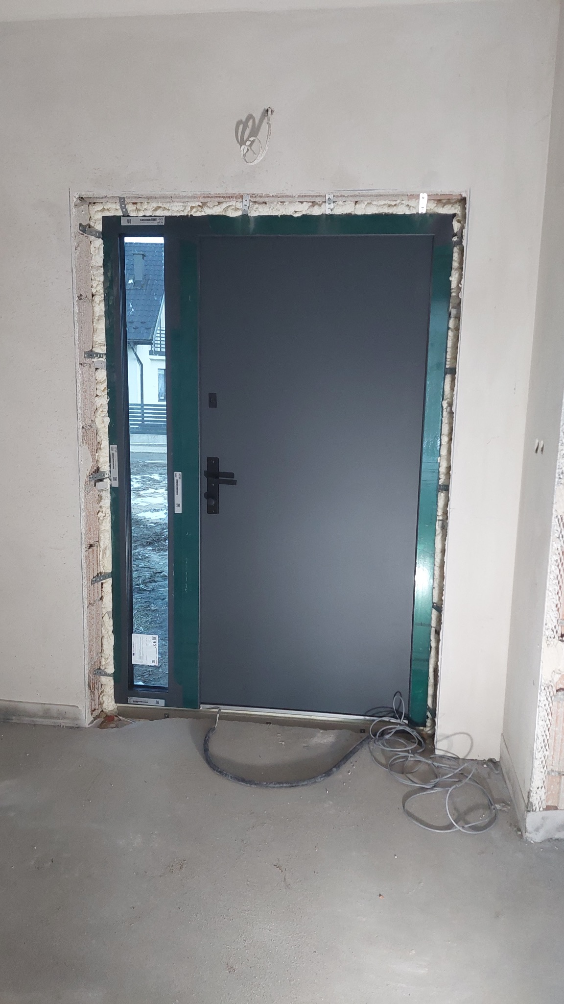 Szara brama wejściowa z ościeżnicą wypełnioną pianką montażową, widok na zewnątrz przez szybę w ościeżnicy, szary przewód na surowej posadzce.