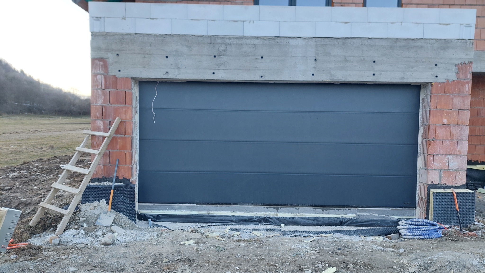 Szara brama garażowa segmentowa w trakcie montażu w surowym otoczeniu budowy. Widoczne cegły, drabina i materiały izolacyjne.