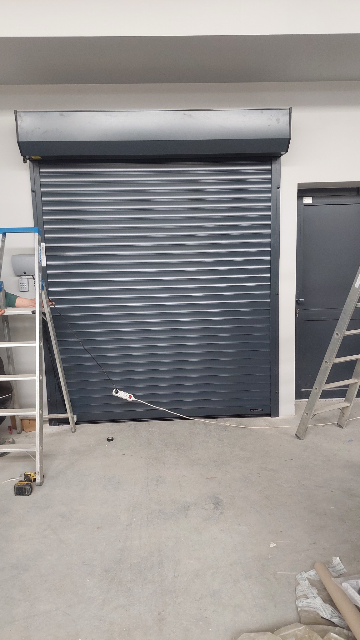 Zamknięta, szara brama rolowana z widocznymi drabinami monterskimi w Wielogłowach. Widoczny kabel zasilający i gniazdko elektryczne na podłodze.