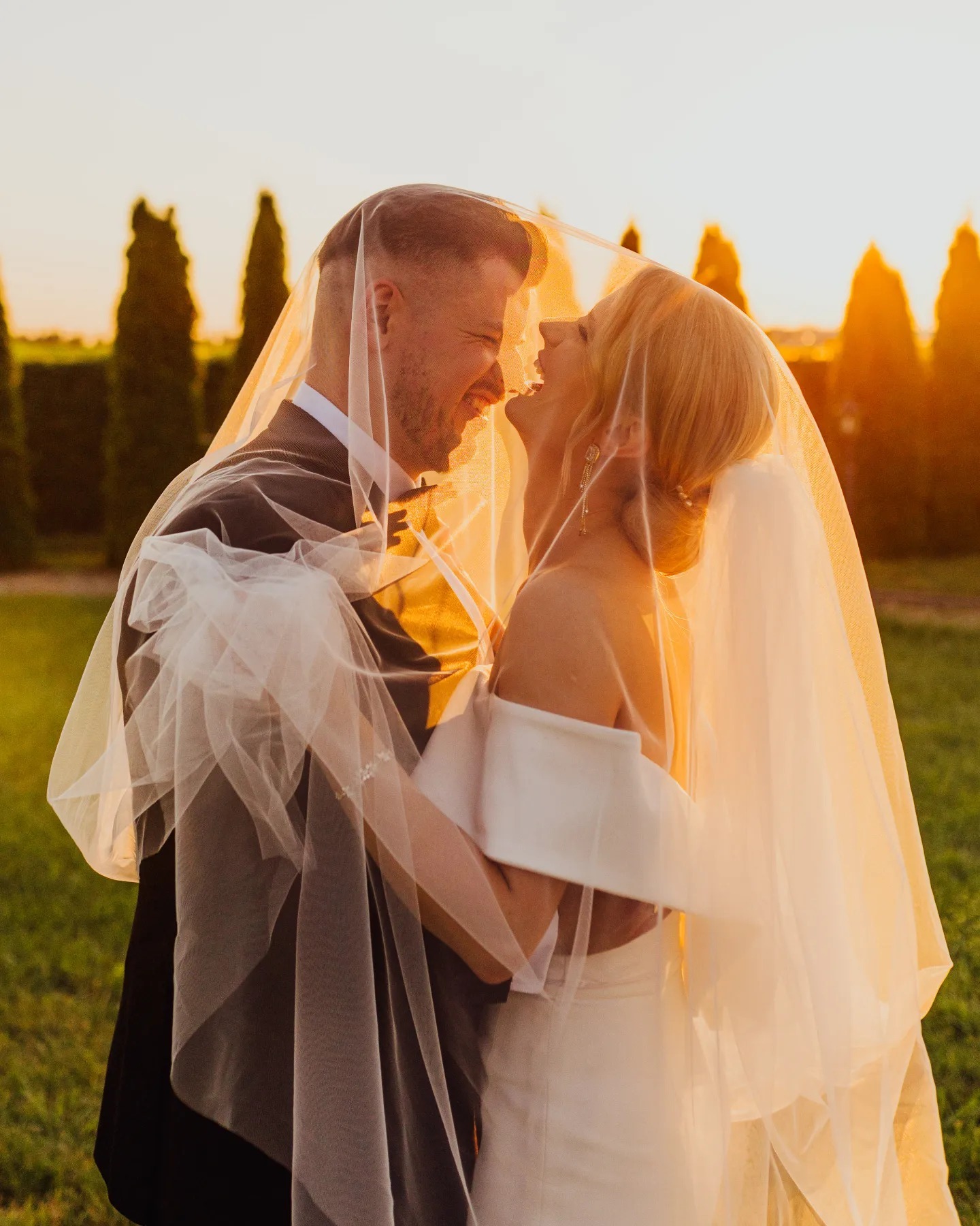 Uśmiechnięta para młoda, częściowo zasłonięta welonem, w promieniach zachodzącego słońca na tle zieleni i wysokich drzew.