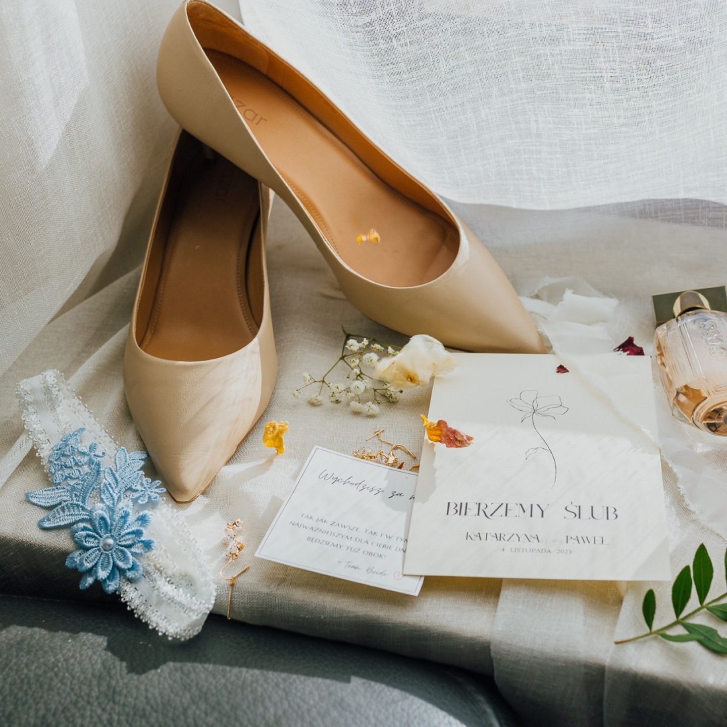 Beżowe damskie czółenka, podwiązka z niebieską koronką, zaproszenie ślubne Katarzyny i Pawła na 4 listopada 2023, perfumy i drobne kwiaty na jasnym materiale.