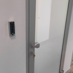 PS-Tech Piotr Skowronek - Szklane drzwi z szarą ramą i kluczem w zamku, obok ściany zamontowany panel dotykowy z klawiaturą numeryczną do kontroli dostępu.
