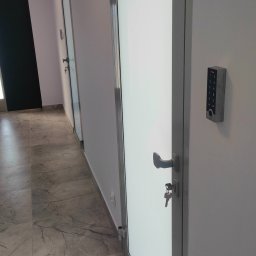 PS-Tech Piotr Skowronek - Białe drzwi z panelem dostępu na kod i kluczem w zamku, widok na korytarz z marmurową podłogą i kolejnymi drzwiami.