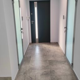 PS-Tech Piotr Skowronek - Wnętrze korytarza z drzwiami wejściowymi z pionowym oświetleniem LED i dwoma szklanymi drzwiami po bokach, szare płytki podłogowe imitujące marmur.