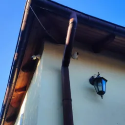 Dwie kamery monitoringu IP zamontowane na elewacji domu, widoczna rynna spustowa i lampa zewnętrzna w stylu retro.