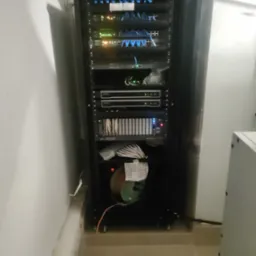 Czarna szafa RACK z widocznymi panelami krosowniczymi, routerami, przełącznikami i okablowaniem, zainstalowana w pomieszczeniu technicznym z białymi ścianami i beżową podłogą.