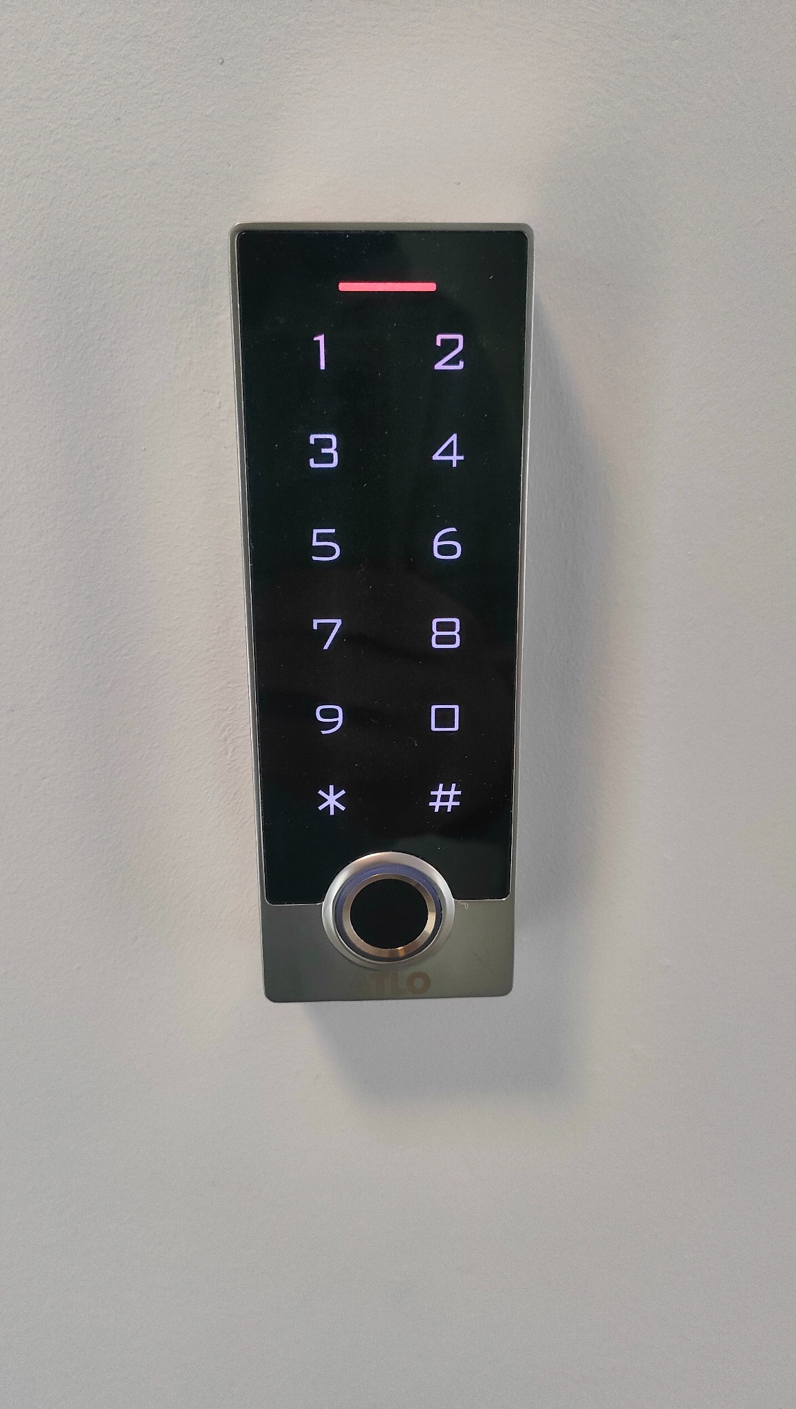 Panel dotykowy kontroli dostępu z czytnikiem linii papilarnych i klawiaturą numeryczną, zamontowany na białej ścianie.