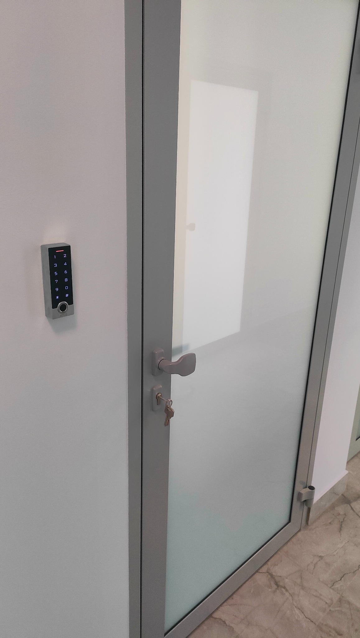 Szklane drzwi z szarą ramą i kluczem w zamku, obok ściany zamontowany panel dotykowy z klawiaturą numeryczną do kontroli dostępu.