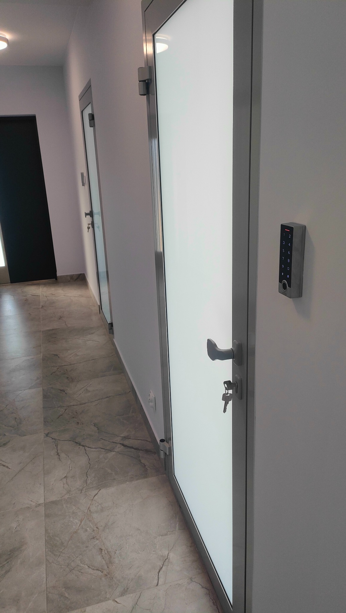 Białe drzwi z panelem dostępu na kod i kluczem w zamku, widok na korytarz z marmurową podłogą i kolejnymi drzwiami.