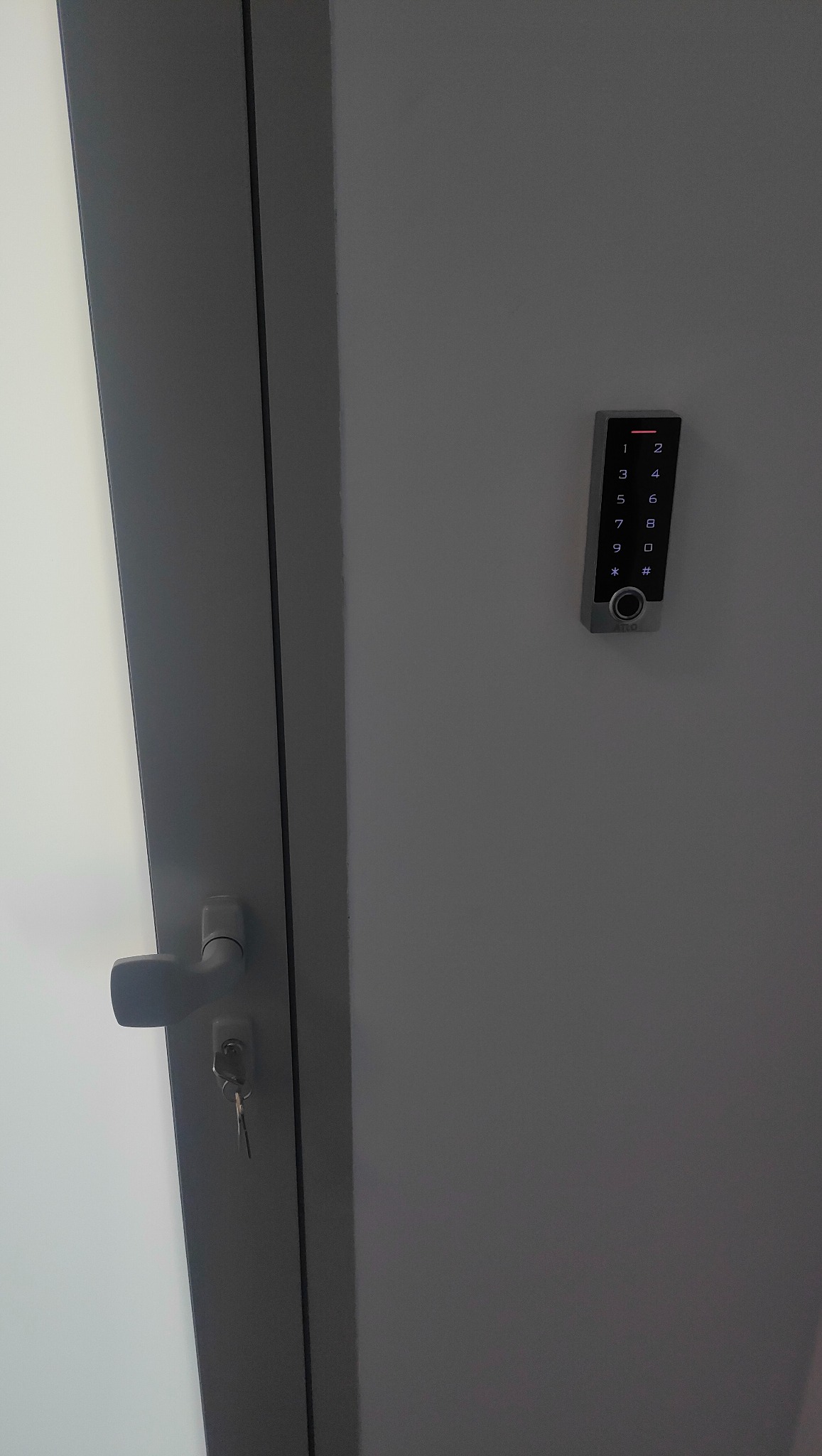 Szary panel z klawiaturą numeryczną i czytnikiem zbliżeniowym zamontowany na drzwiach, obok klamki z kluczem w zamku.