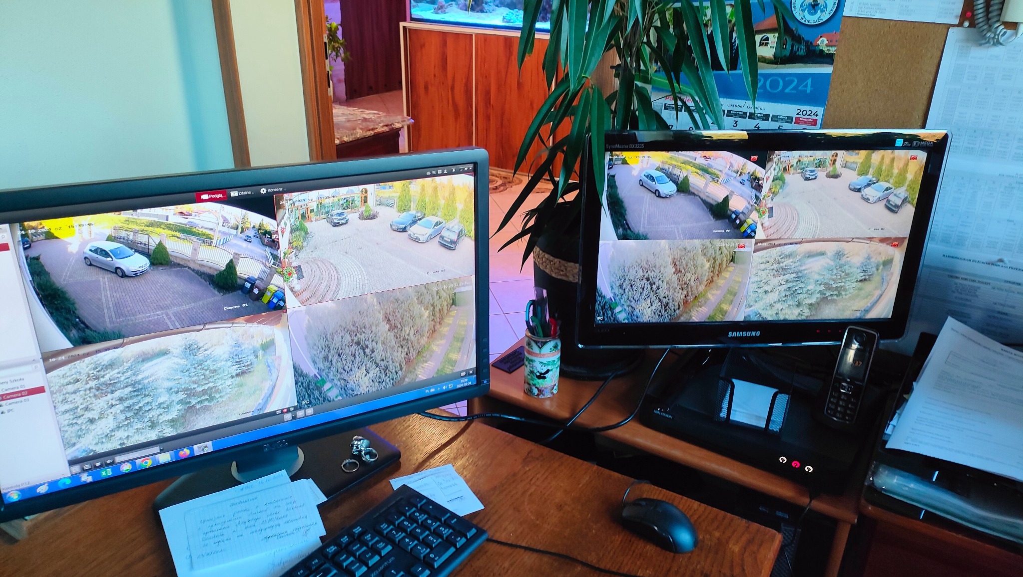 Dwa monitory wyświetlają obraz z kamer monitoringu: na jednym widok na podjazd z zaparkowanymi samochodami, na drugim widok z kamery na zieleń. Monitory stoją na biurku, obok klawiatura, myszka...