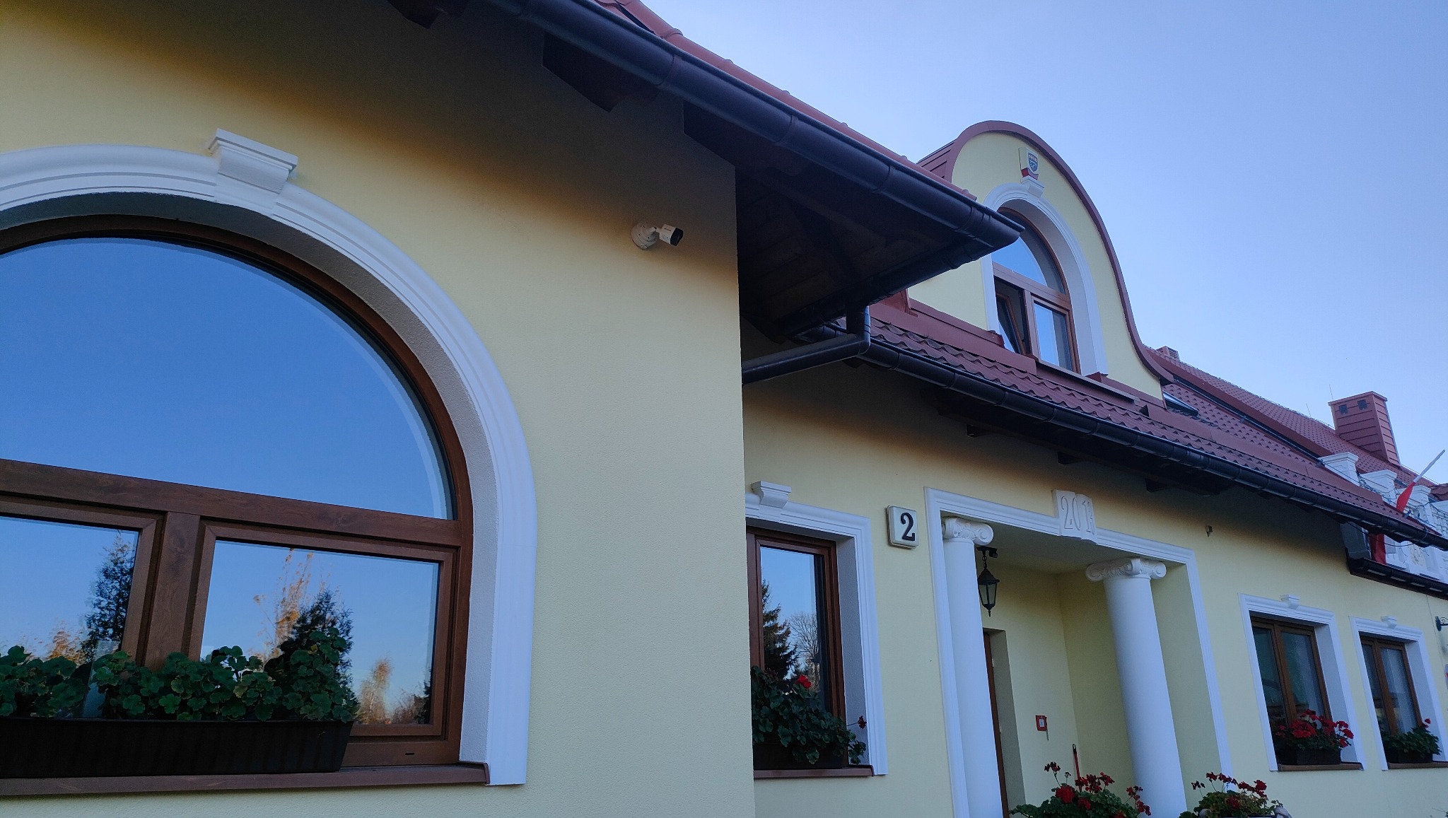 Kamera monitoringu zewnętrznego zamontowana na elewacji budynku z żółtą fasadą, widoczna pod rynną, obok okna z półokrągłym łukiem.