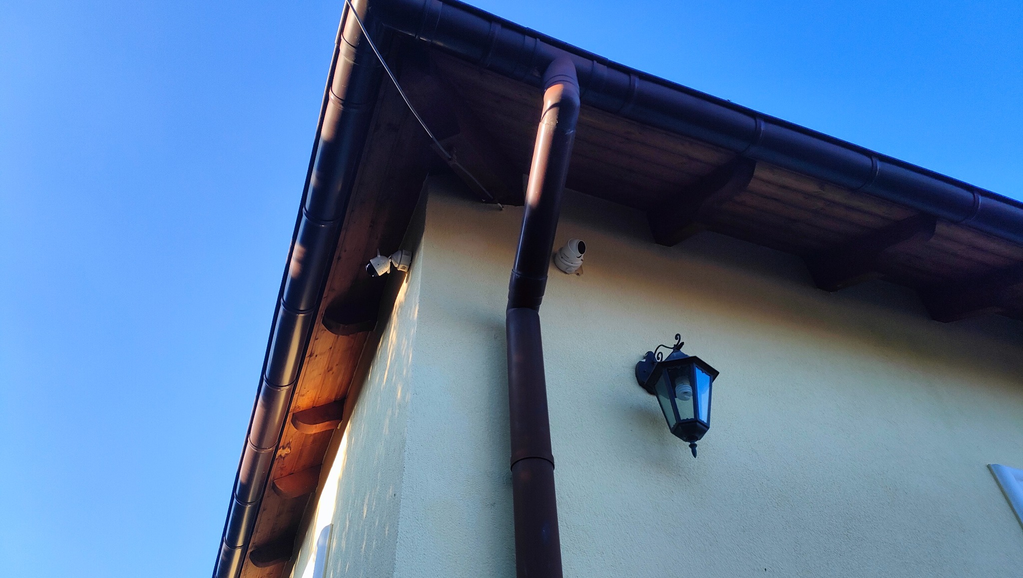Dwie kamery monitoringu IP zamontowane na elewacji domu, widoczna rynna spustowa i lampa zewnętrzna w stylu retro.