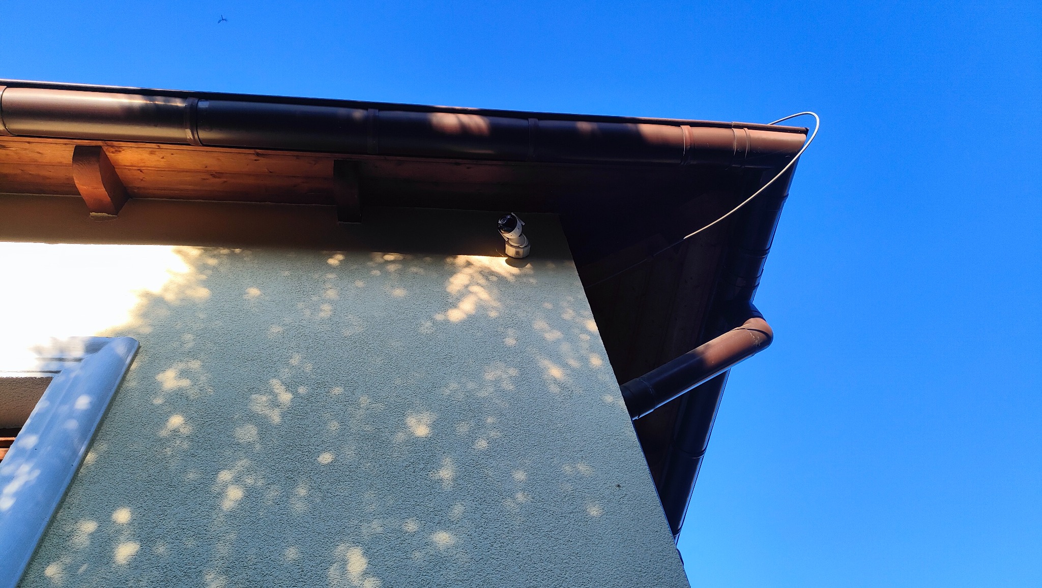 Kamera IP zamontowana na elewacji budynku pod brązowym orynnowaniem, widoczne cienie liści na ścianie i przewód zasilający.