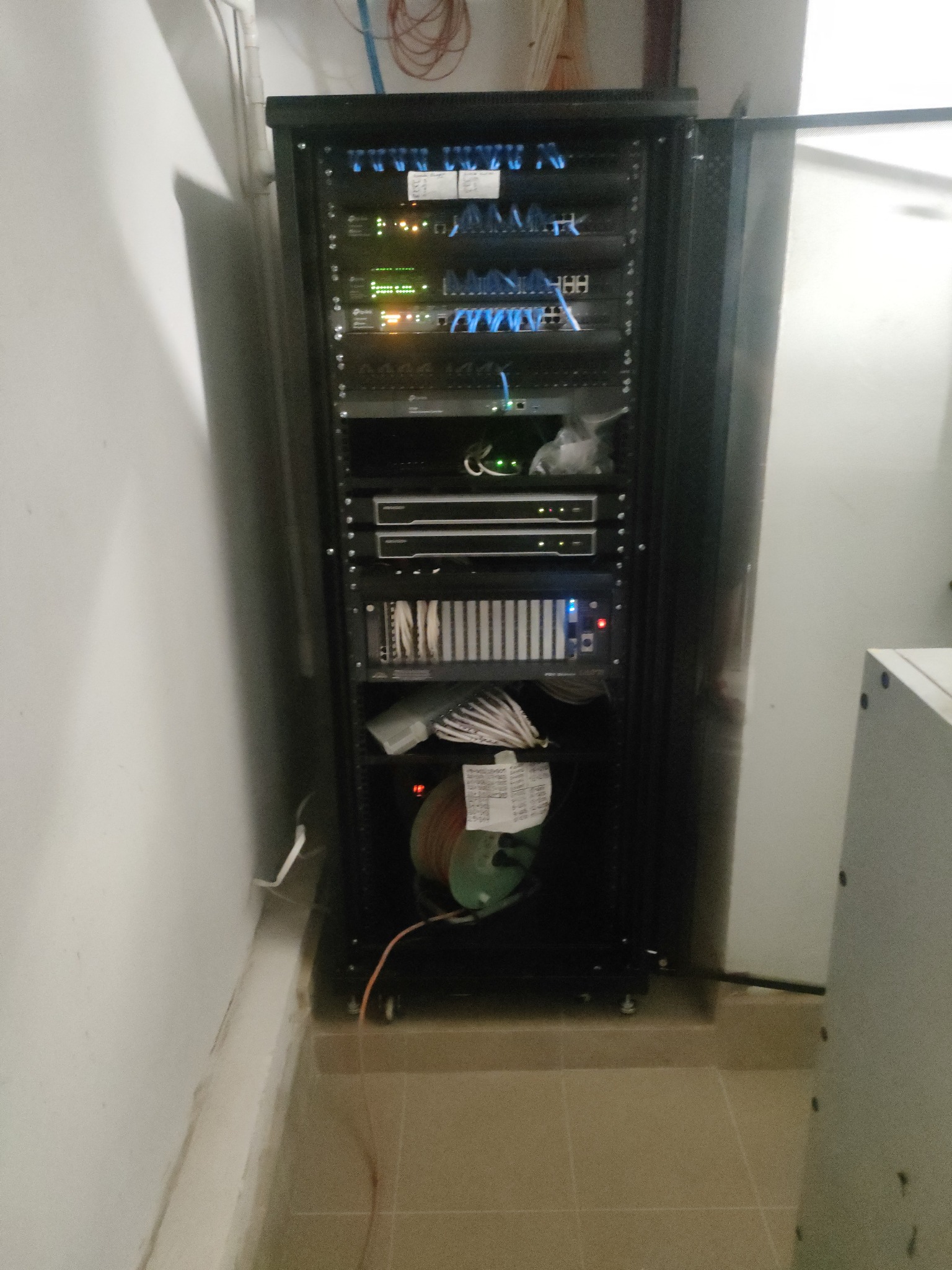 Czarna szafa RACK z widocznymi panelami krosowniczymi, routerami, przełącznikami i okablowaniem, zainstalowana w pomieszczeniu technicznym z białymi ścianami i beżową podłogą.