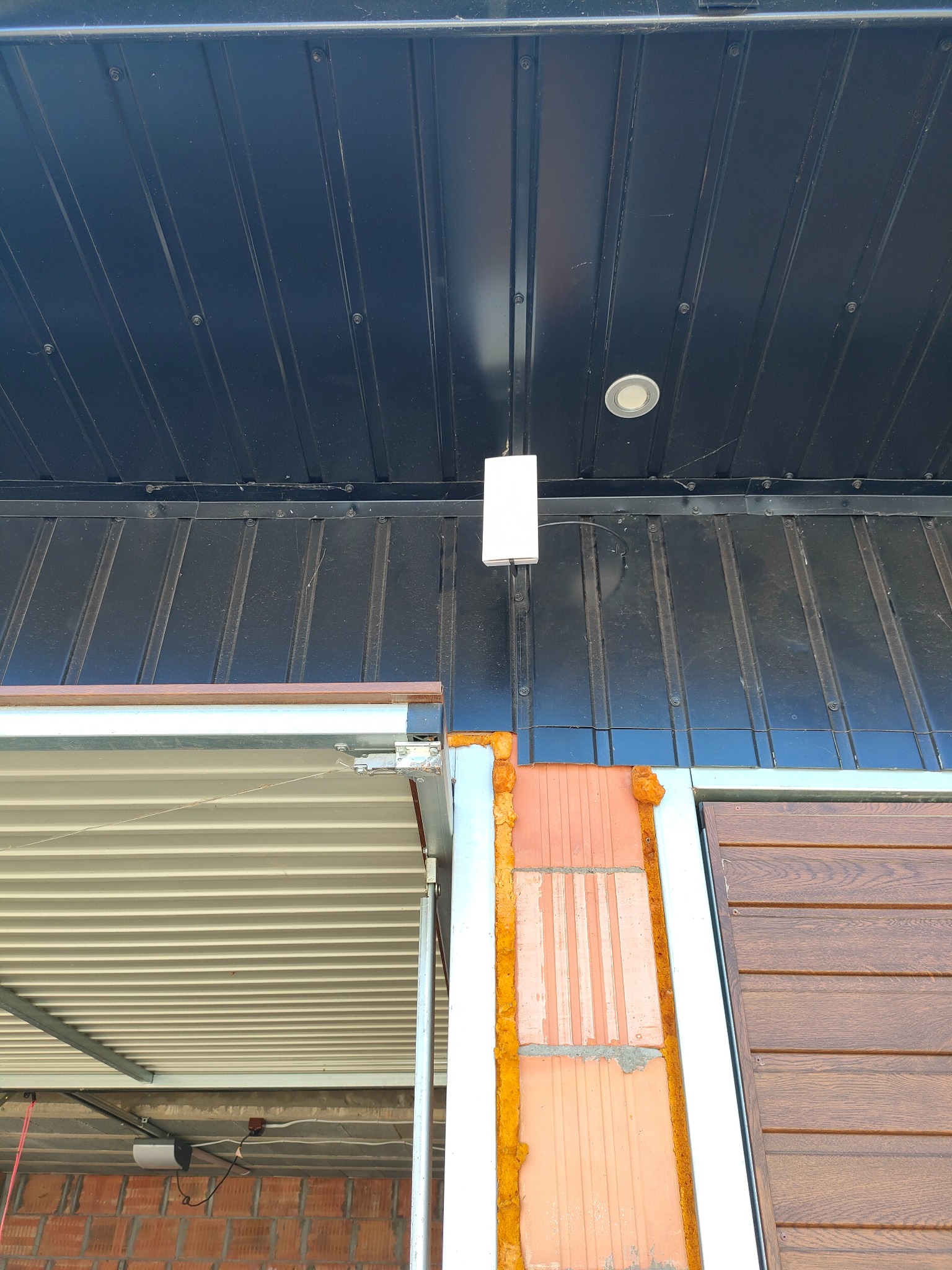 Biały punkt dostępowy zamontowany na czarnej blasze falistej sufitu, widoczne przewody, fragment ściany z cegły i pianki montażowej oraz brama garażowa.