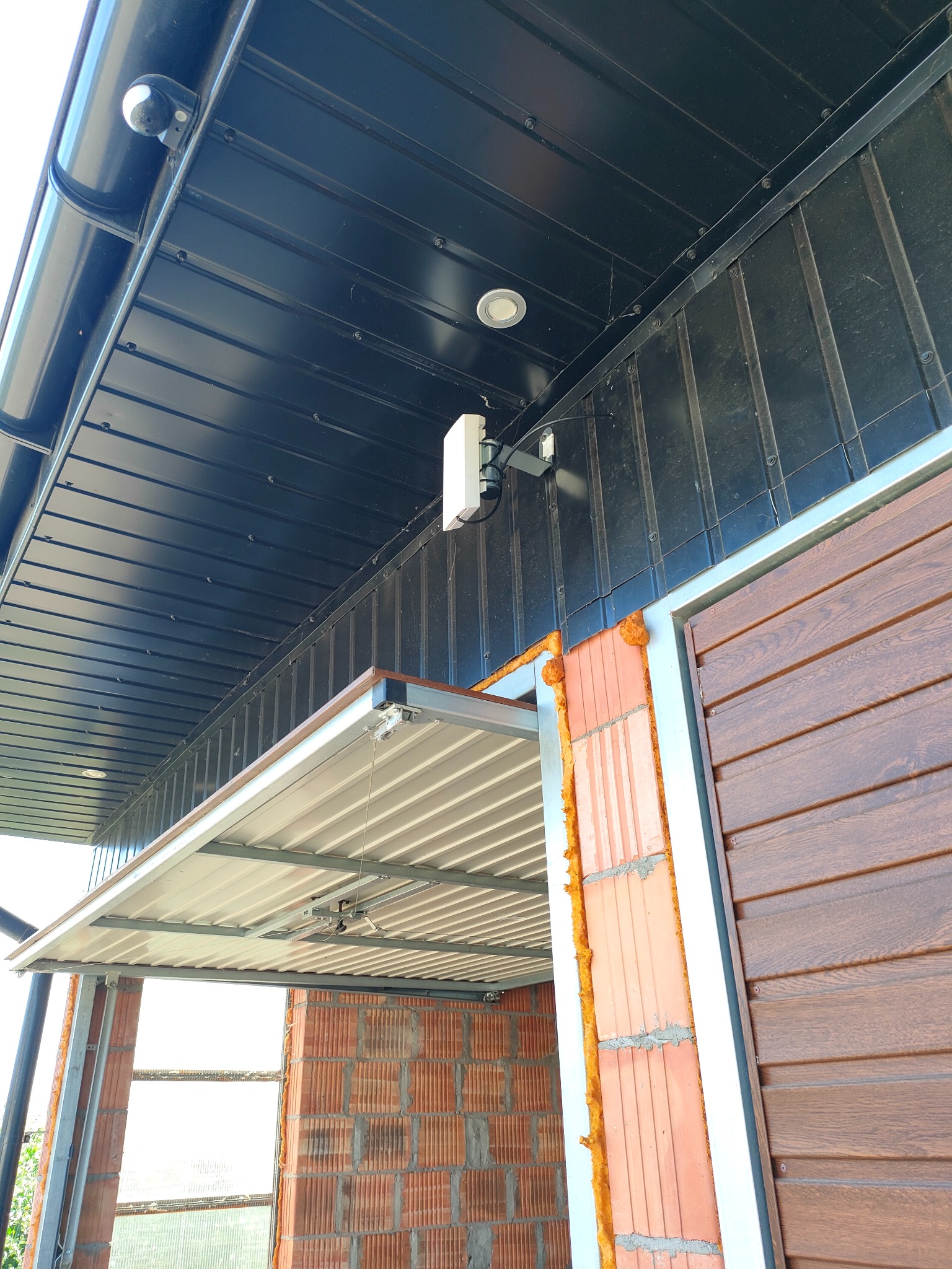 Zewnętrzna kamera monitoringu zamontowana pod czarnym, metalowym dachem, obok białego punktu dostępowego WiFi, z widoczną instalacją elektryczną i fragmentem ściany z brązową okładziną imitującą...