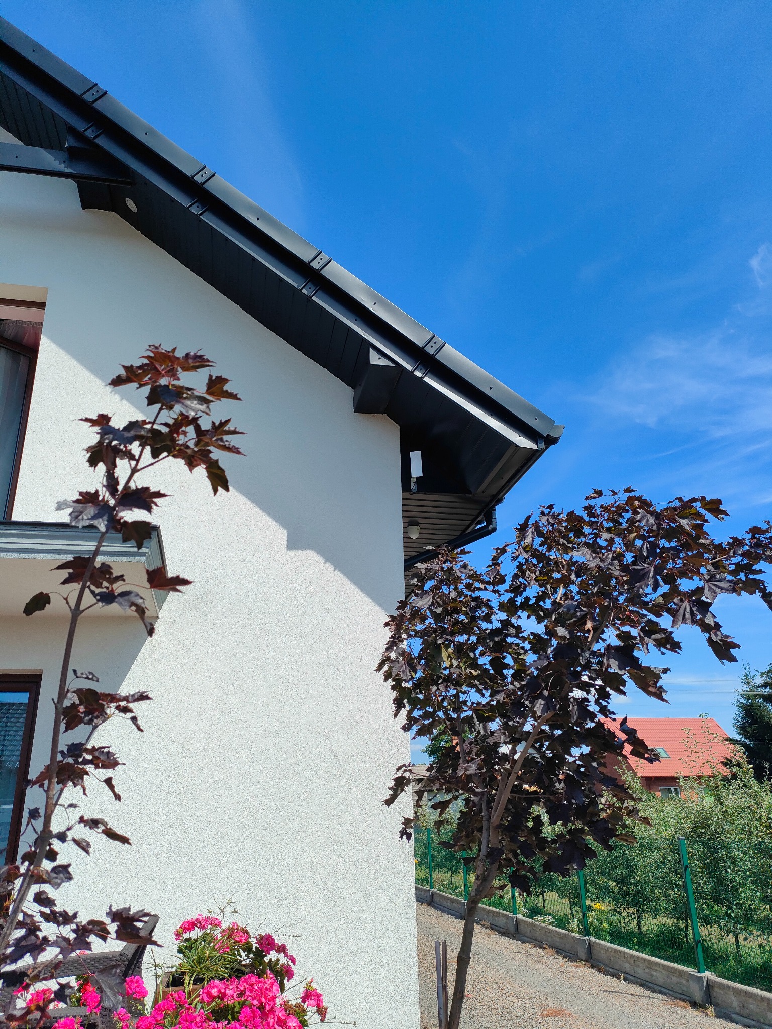 Biała ściana domu z zamontowanym, dyskretnym, białym czujnikiem ruchu pod okapem, widocznym między dwoma drzewami o purpurowych liściach na tle błękitnego nieba.