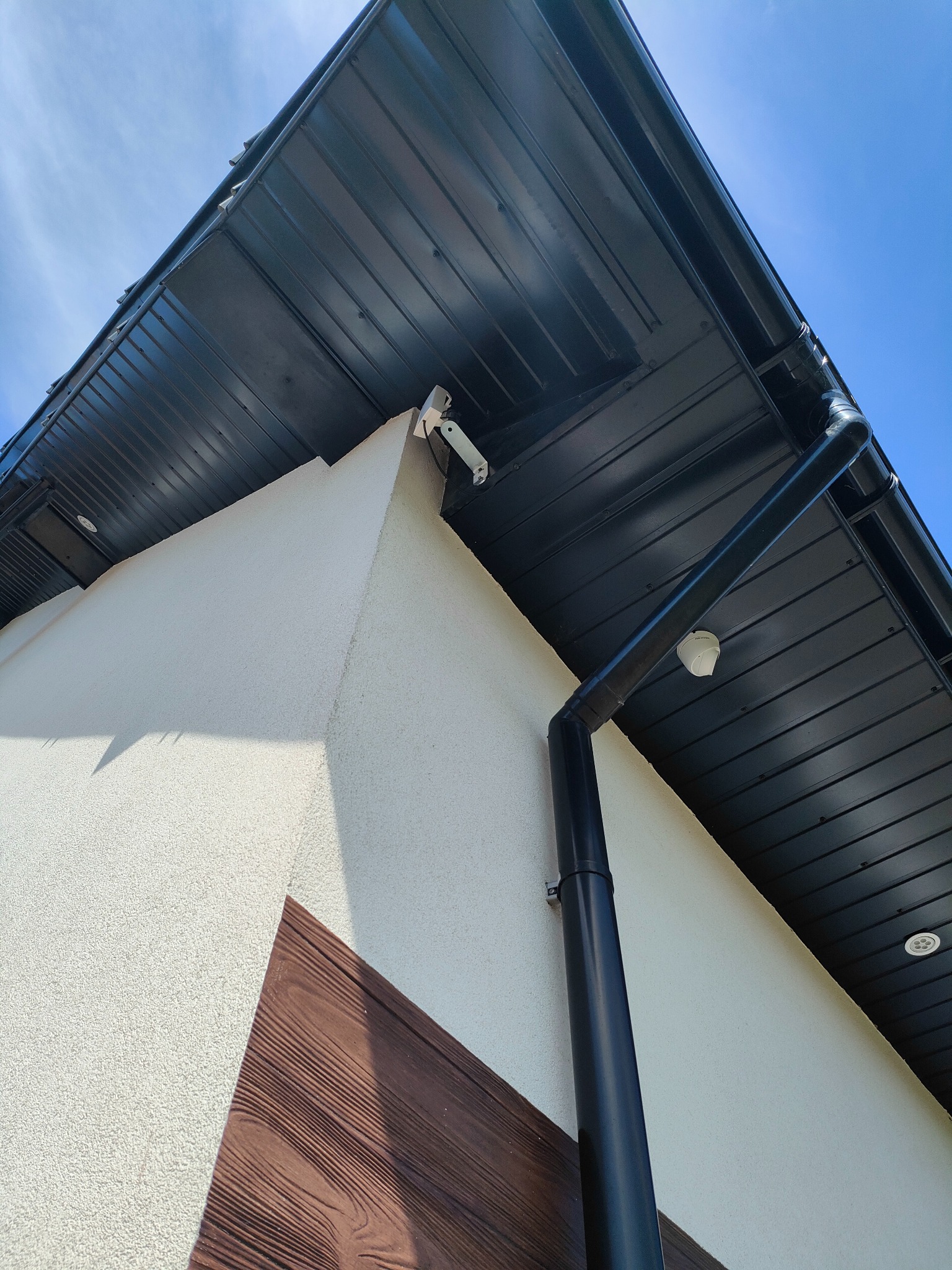 Zewnętrzna kamera monitoringu IP zamontowana pod okapem budynku z ciemną blachodachówką i rynną spustową, widoczna z dołu pod kątem, na tle błękitnego nieba.