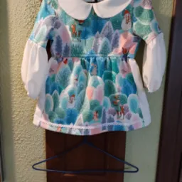 Sukienka dziecięca z motywem zimowego lasu i zwierząt, wykończona białym kołnierzykiem i koronką, wisząca na wieszaku.