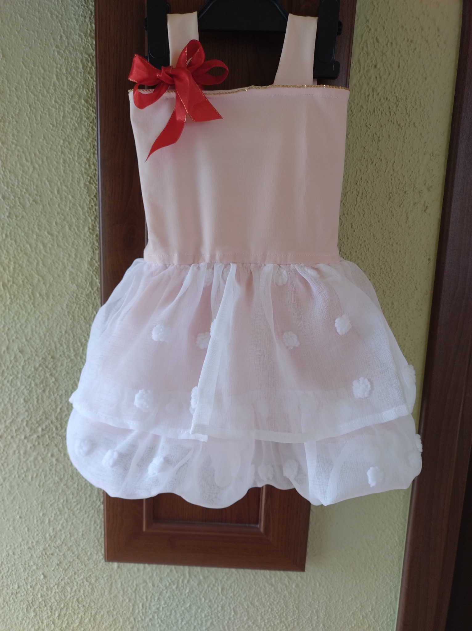 Różowa sukienka dziewczęca z tiulowym dołem w białe kropki, ozdobiona czerwoną kokardą, wisząca na wieszaku na tle ściany i framugi drzwi.