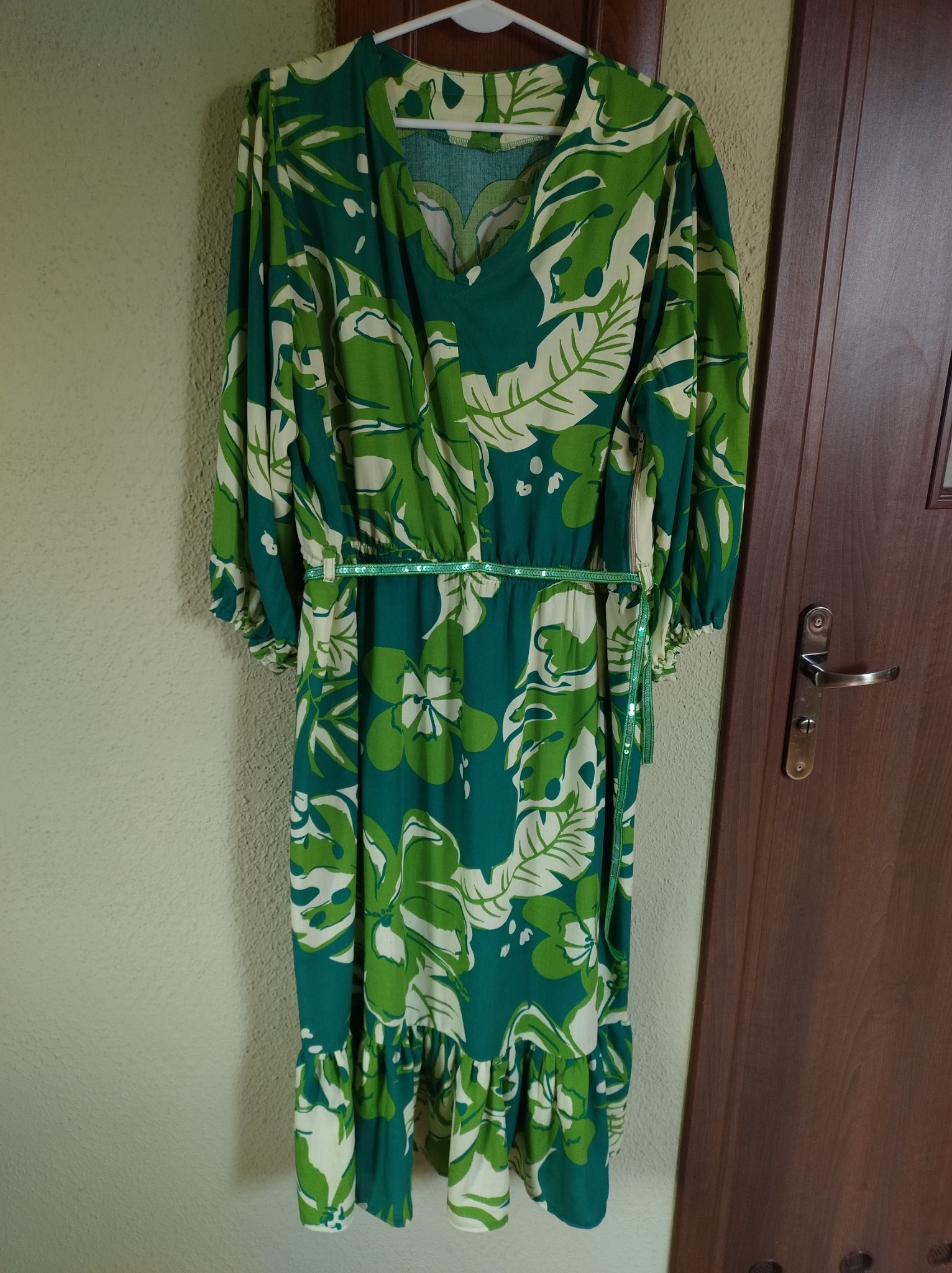 Długa, letnia sukienka w odcieniach zieleni i bieli, z motywem roślinnym, wisząca na wieszaku obok drewnianych drzwi.