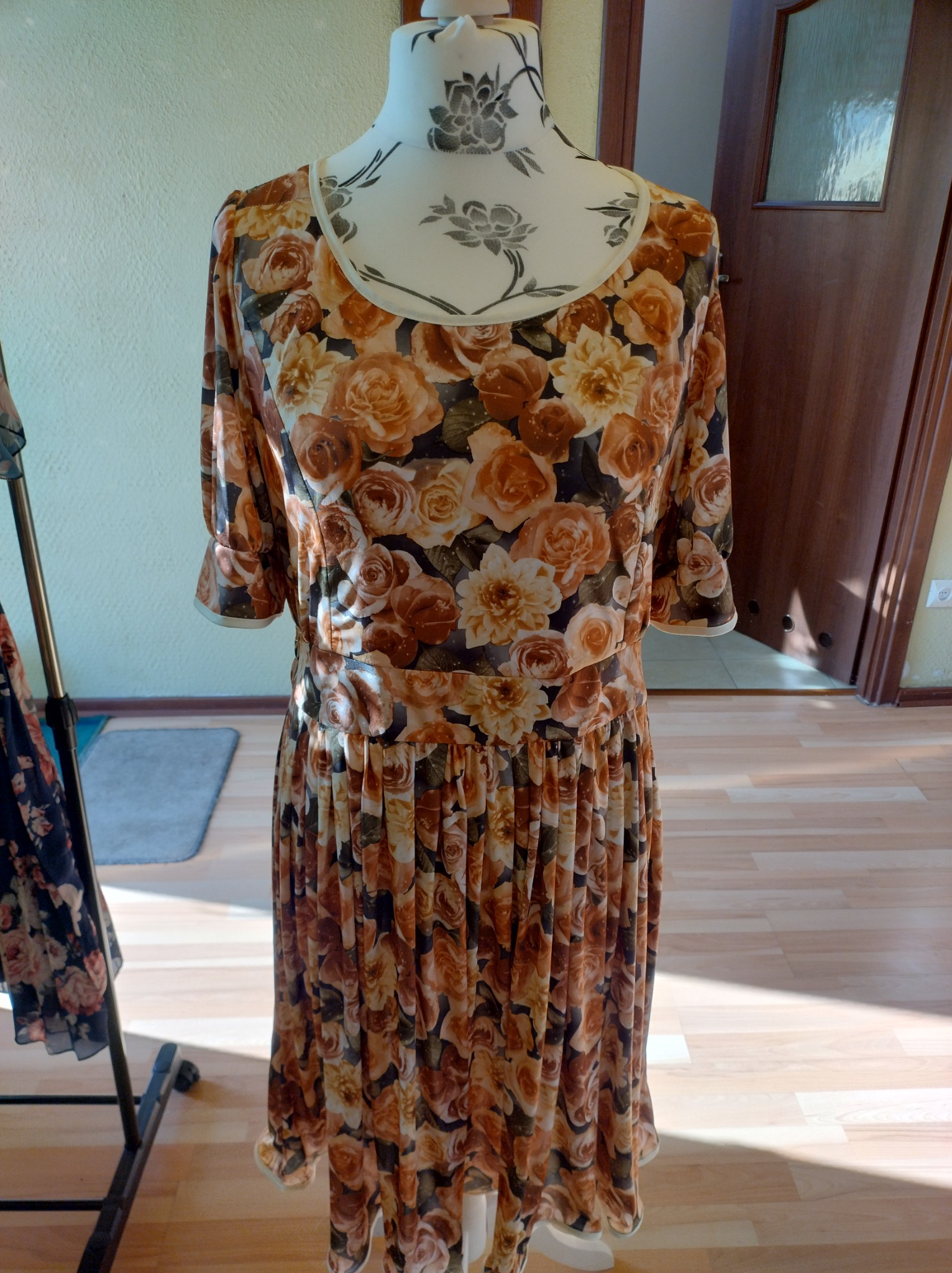 Sukienka damska w kwiaty, w odcieniach brązu i beżu, na manekinie krawieckim z nadrukiem kwiatowym, w tle drewniane drzwi i wieszak na ubrania.