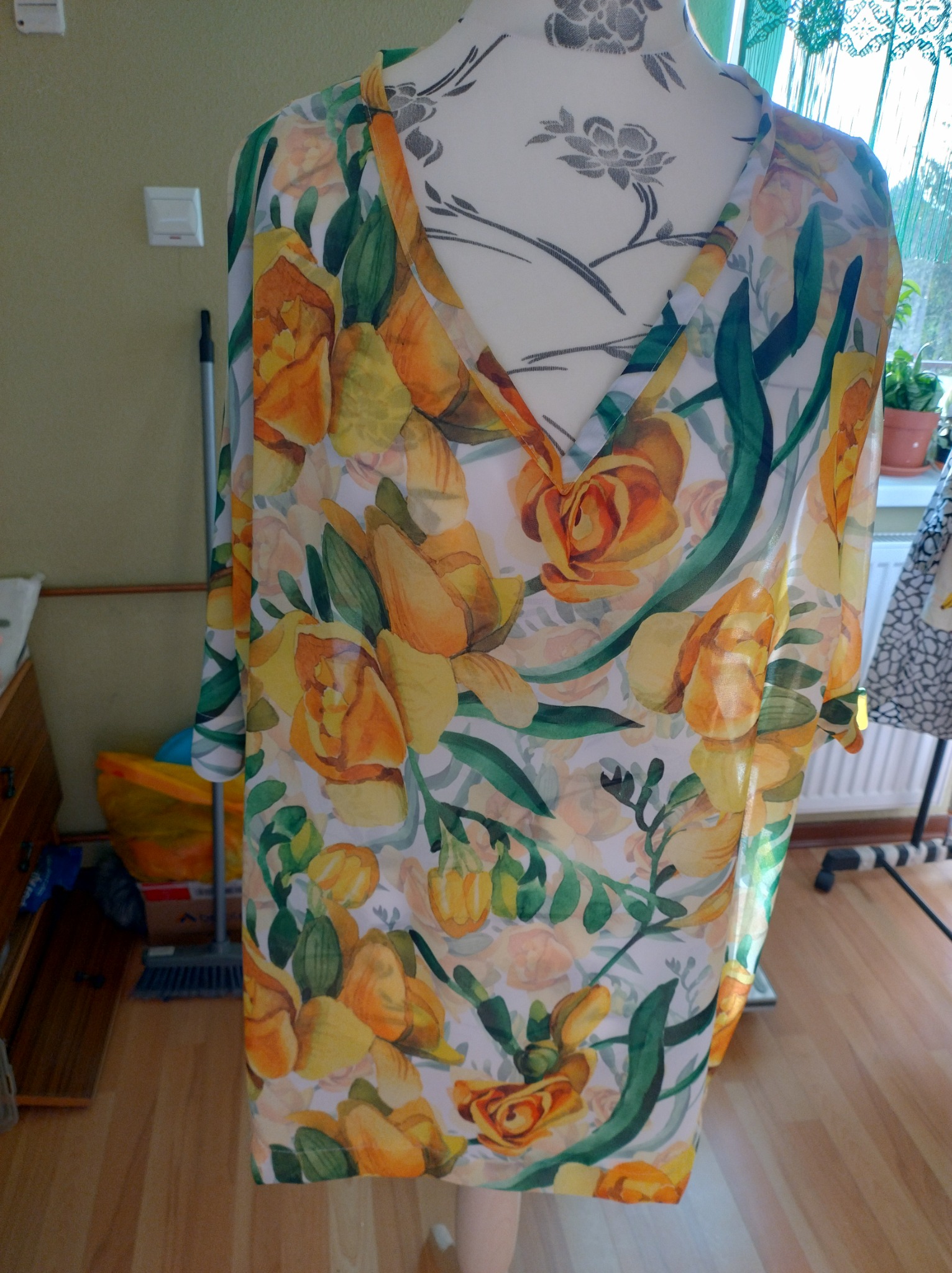 Damska tunika z delikatnej, prześwitującej tkaniny w kwiatowy wzór, zawieszona na manekinie krawieckim w domowym wnętrzu.