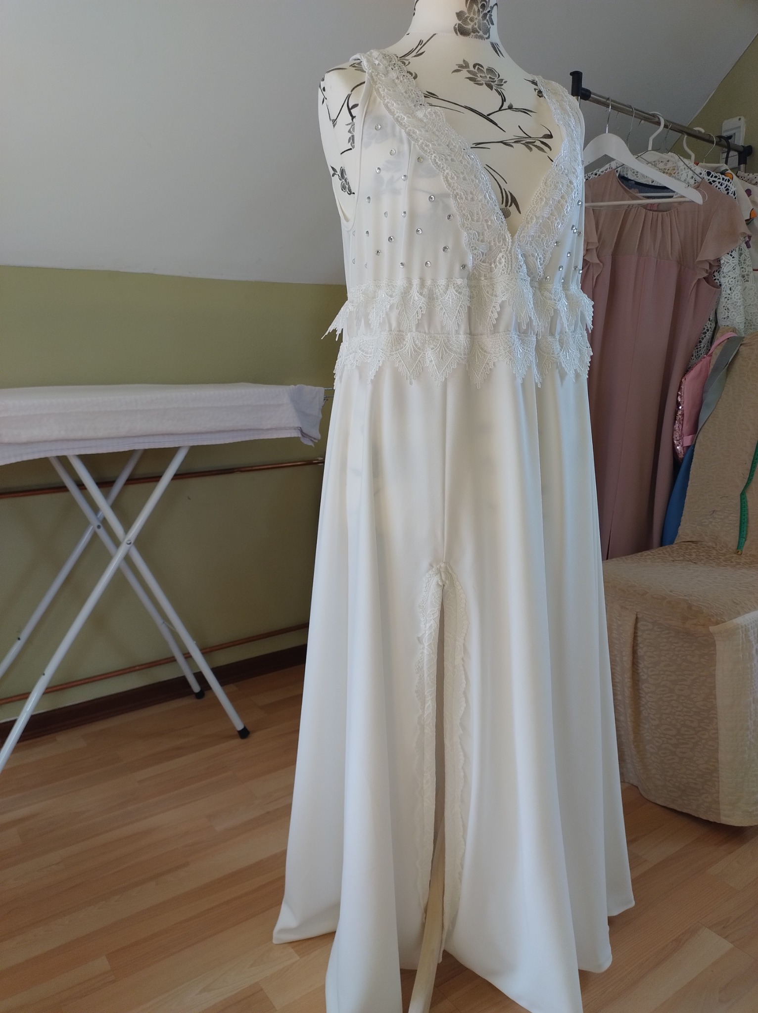 Biała suknia z koronkowymi zdobieniami i rozcięciem na manekinie, w tle deska do prasowania i inne ubrania na wieszaku.