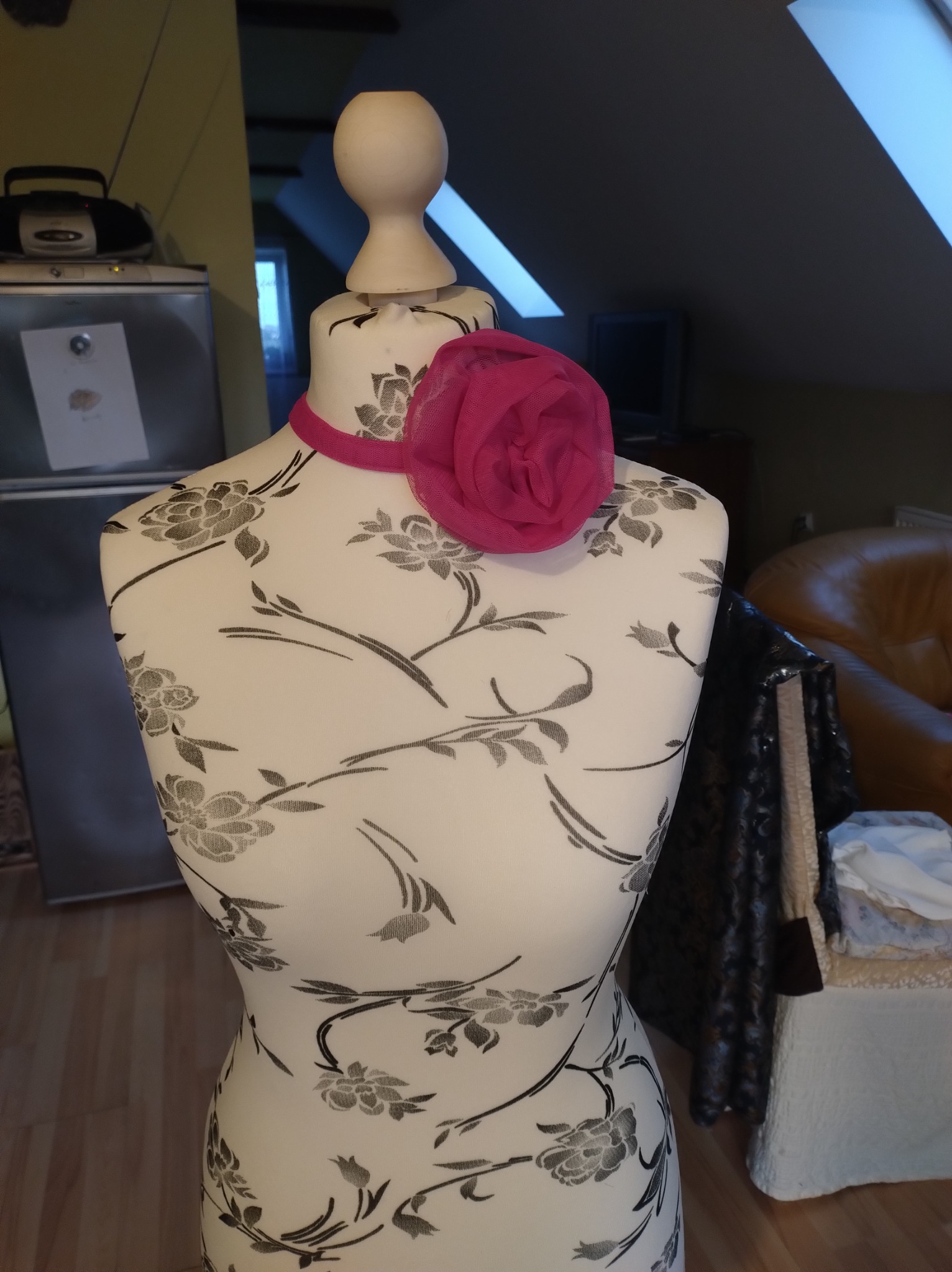 Manekin krawiecki w biało-czarne kwiaty z różową ozdobą na szyi, ustawiony w pomieszczeniu z oknami dachowymi i widocznym sprzętem AGD w tle.