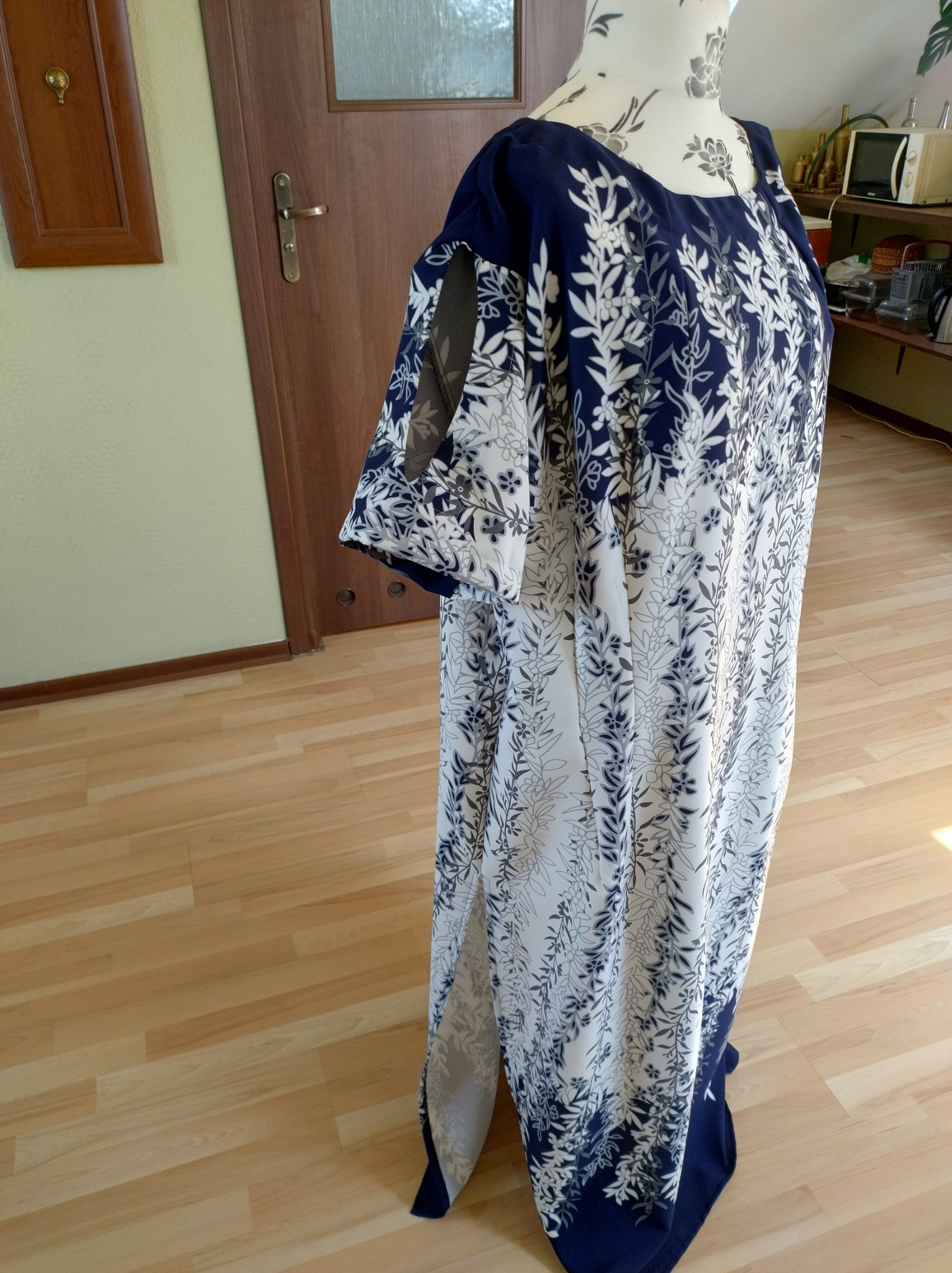 Długa, zwiewna suknia w biało-granatowe kwiaty na manekinie krawieckim, z rozcięciem na boku i krótkimi rękawami z siateczkowymi wstawkami, ustawiona na drewnianej podłodze w pomieszczeniu.