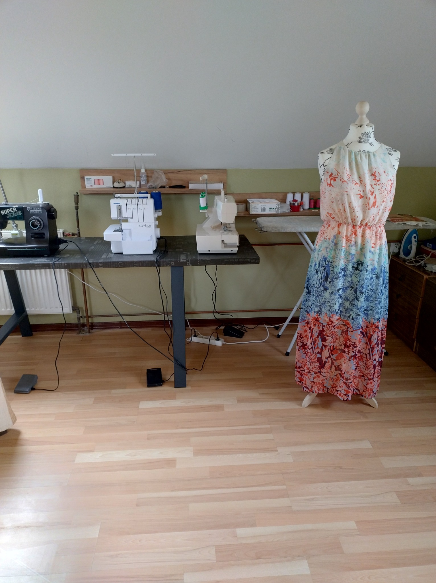 Pracownia krawiecka z manekinem prezentującym zwiewną, wielobarwną sukienkę w kwiaty, maszyny do szycia na stole i deską do prasowania w tle.
