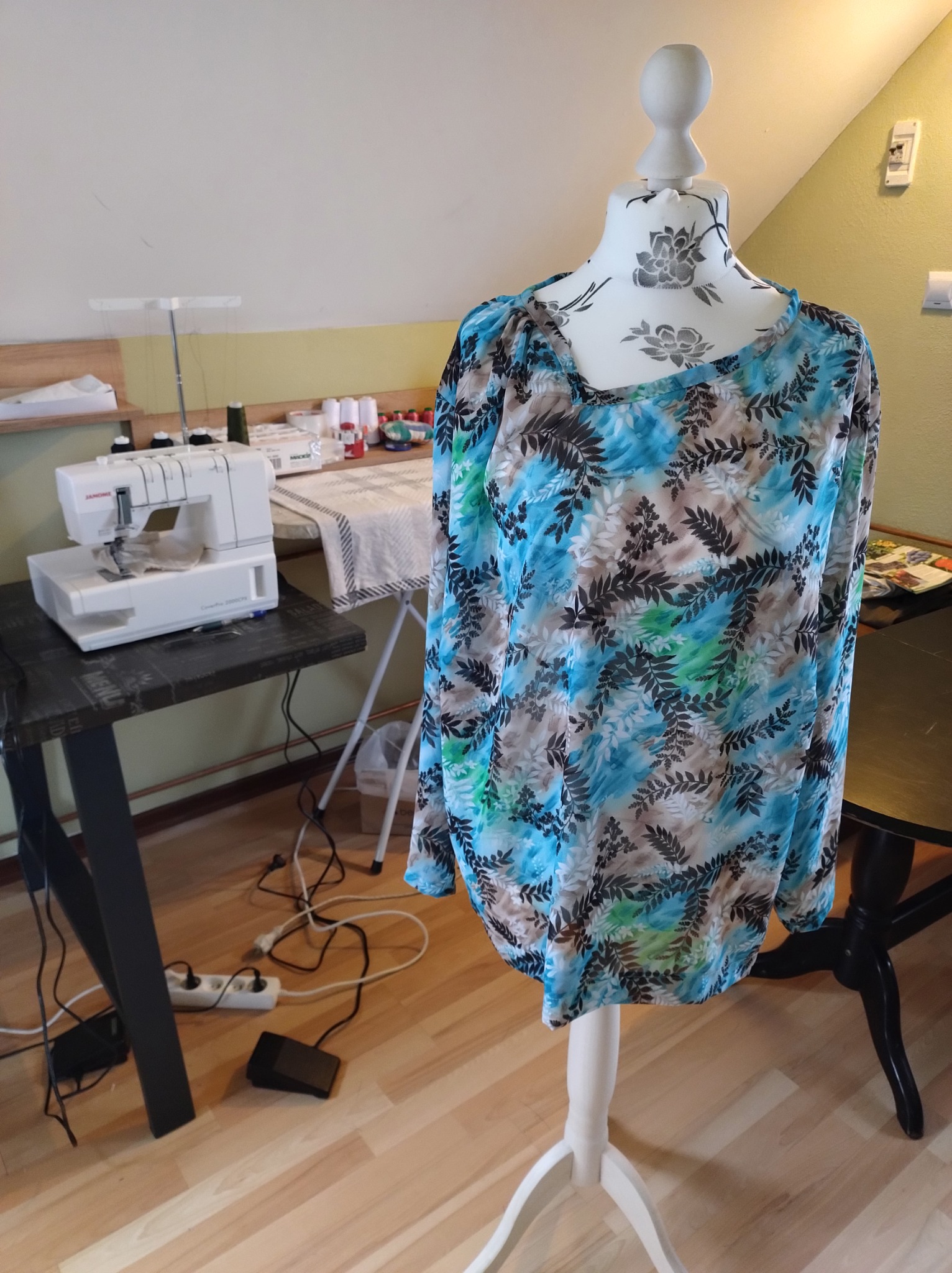 Bluzka z wzorem liści w odcieniach błękitu, brązu i zieleni na manekinie krawieckim, w tle maszyna do szycia, stół z akcesoriami krawieckimi i drewniana podłoga.