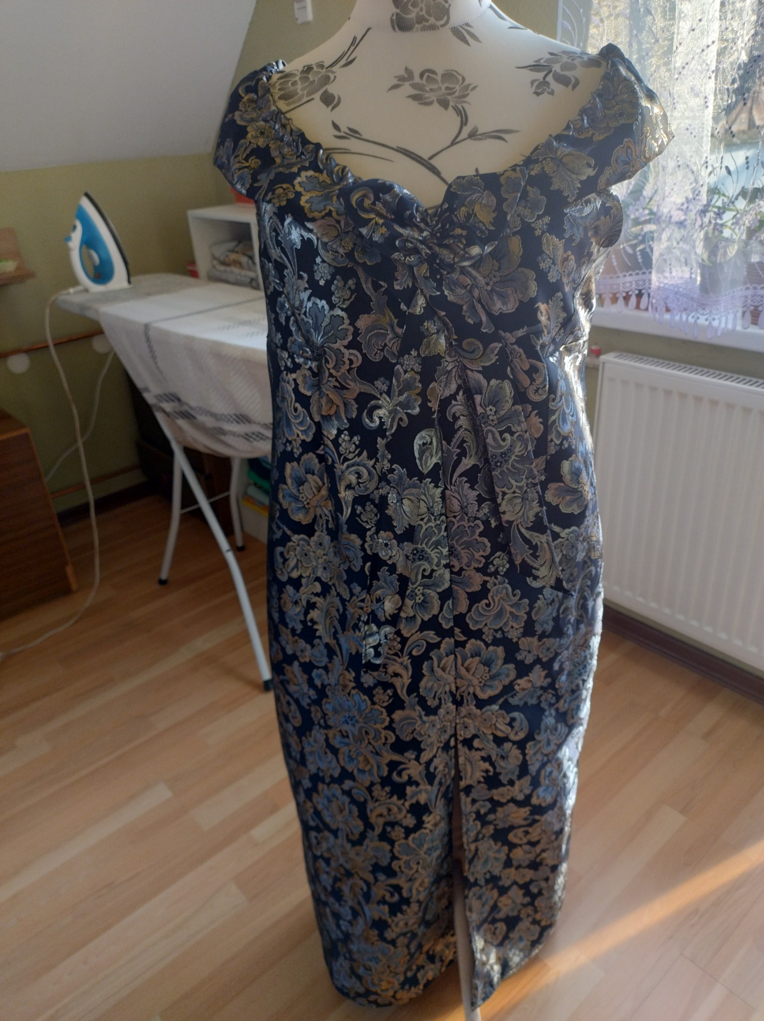 Elegancka suknia wieczorowa z wzorzystego, brokatowego materiału, prezentowana na manekinie krawieckim w pracowni, w tle deska do prasowania i żelazko.
