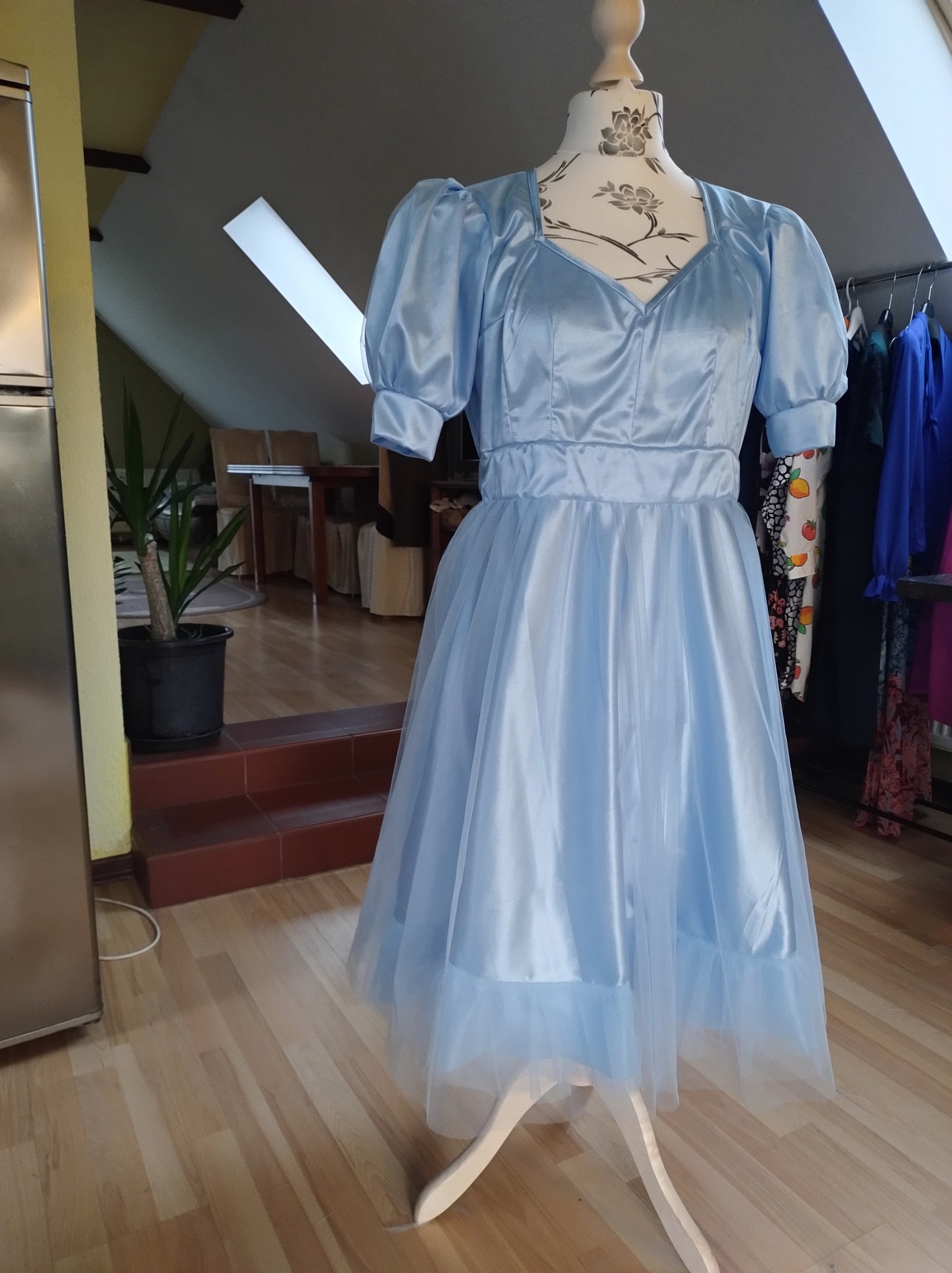 Błękitna, satynowo-tiulowa sukienka z bufkami, prezentowana na manekinie w pracowni krawieckiej z drewnianą podłogą i widocznym fragmentem rośliny doniczkowej.