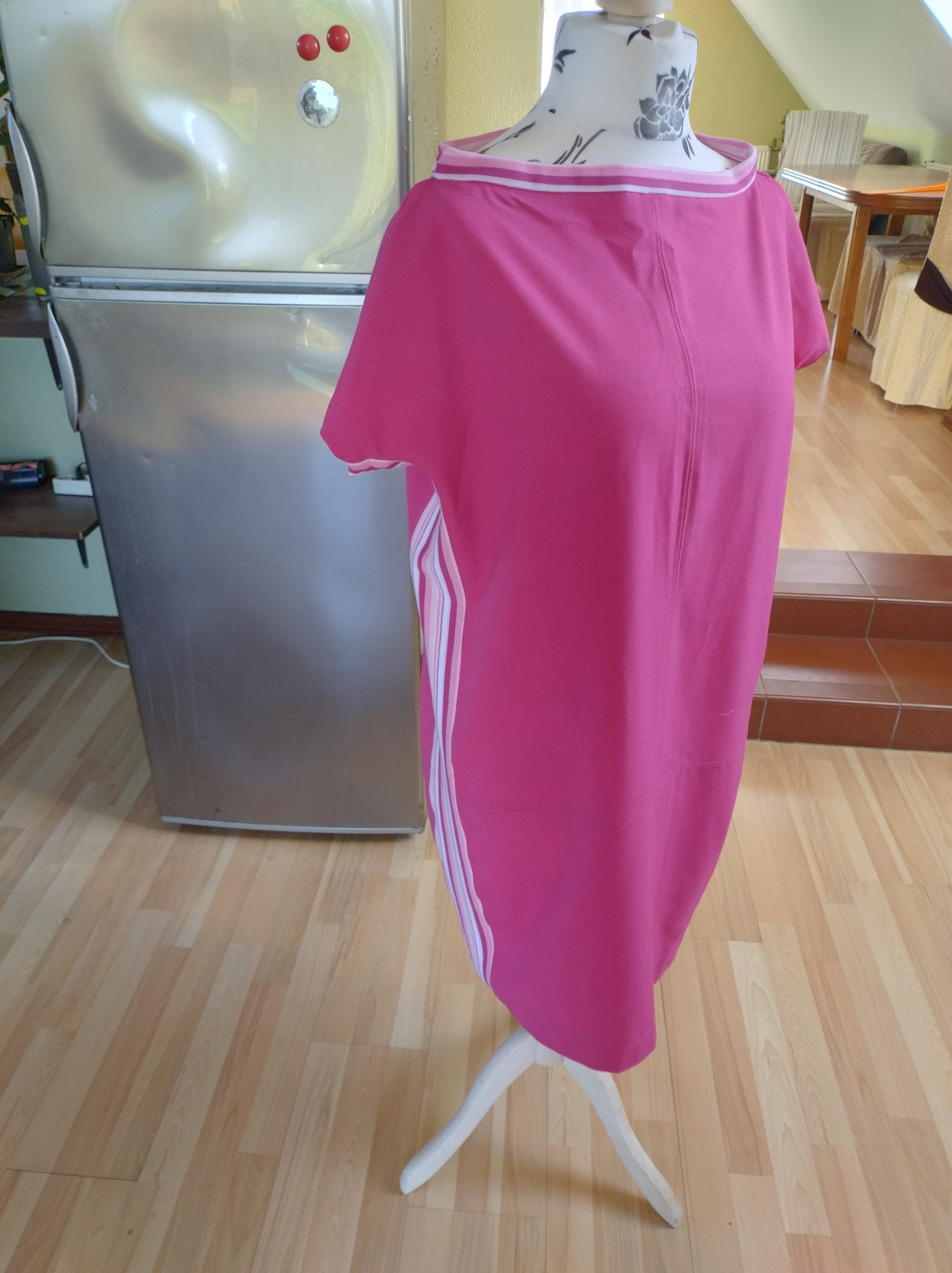 Różowa tunika z białymi paskami po bokach, zaprezentowana na manekinie krawieckim w domowym wnętrzu z widoczną lodówką i drewnianą podłogą.