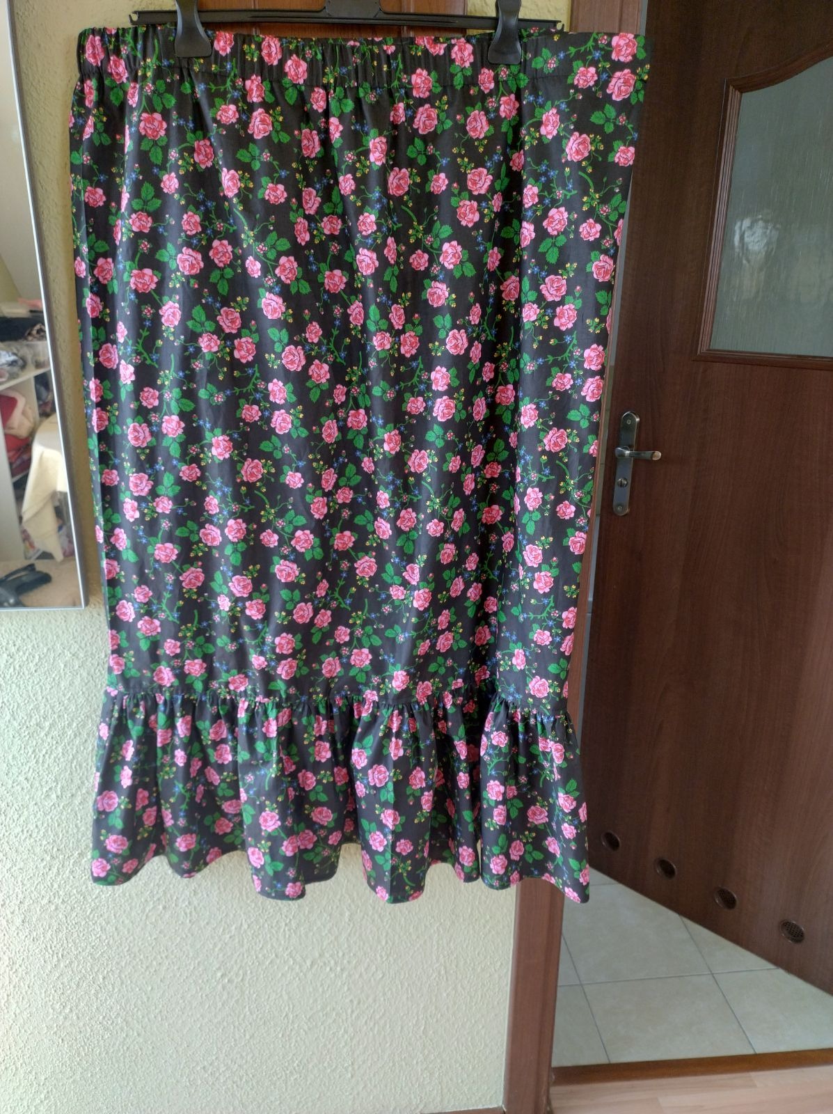 Czarna spódnica z różowym wzorem kwiatowym, wykończona falbaną, wisząca na wieszaku na tle ściany i drzwi.