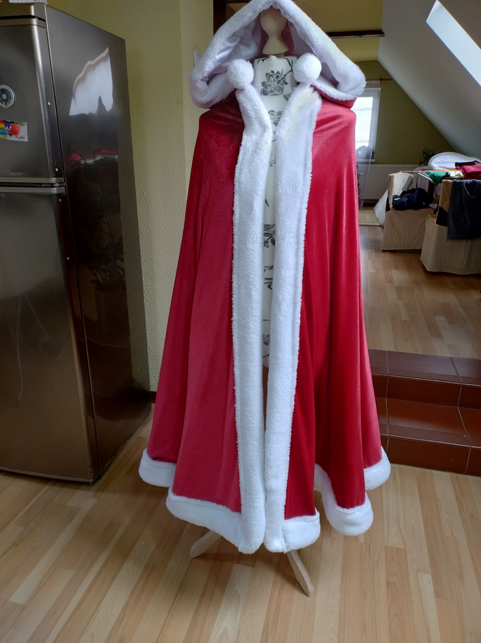 Czerwony, welurowy płaszcz z białym futerkiem, stylizowany na strój Świętego Mikołaja, prezentowany na manekinie w domowym wnętrzu z widoczną lodówką i schodami.