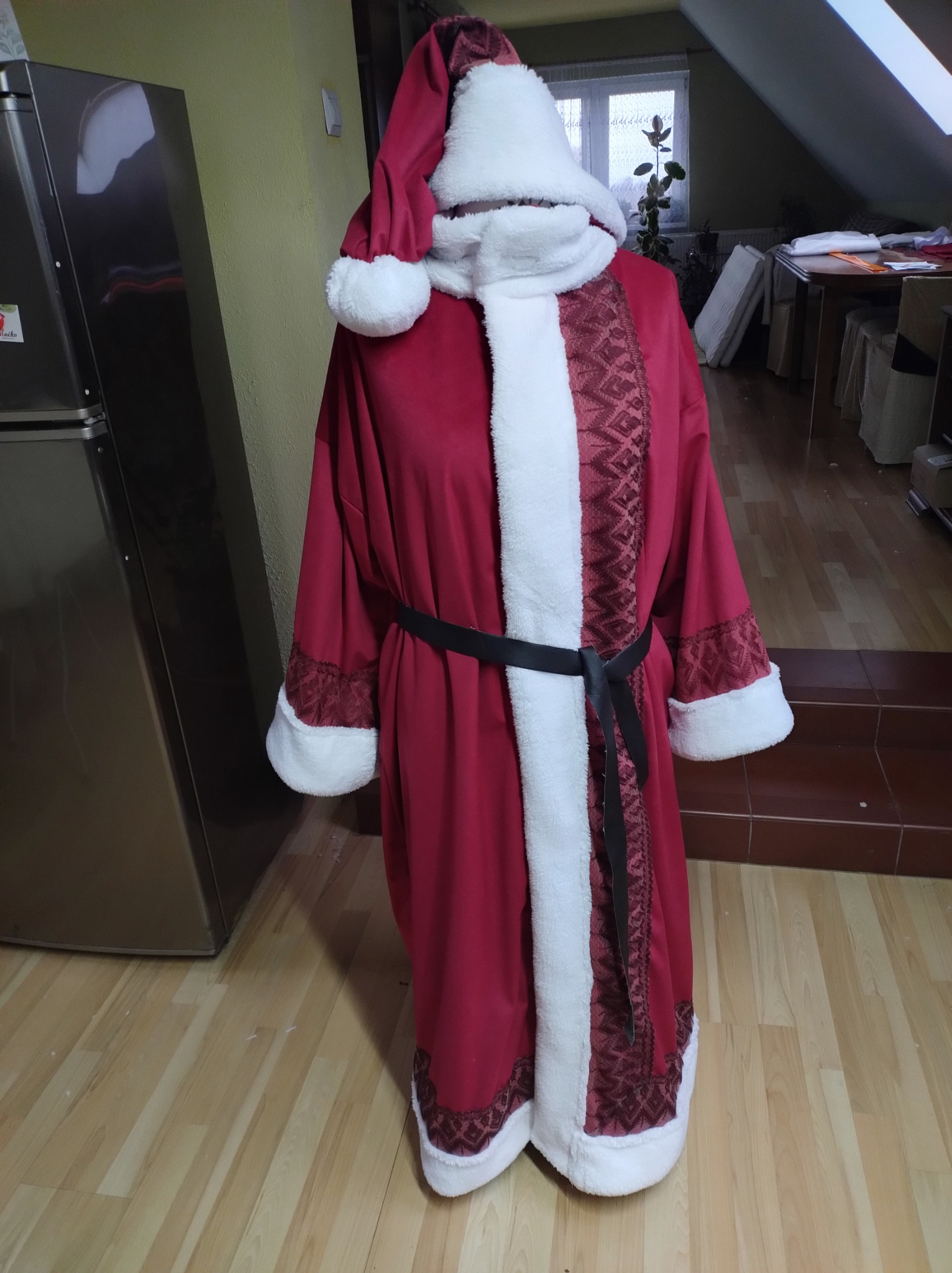 Czerwony strój Świętego Mikołaja z białym futerkiem i czarnym paskiem, prezentowany na manekinie w domowym wnętrzu z widoczną lodówką i oknem.