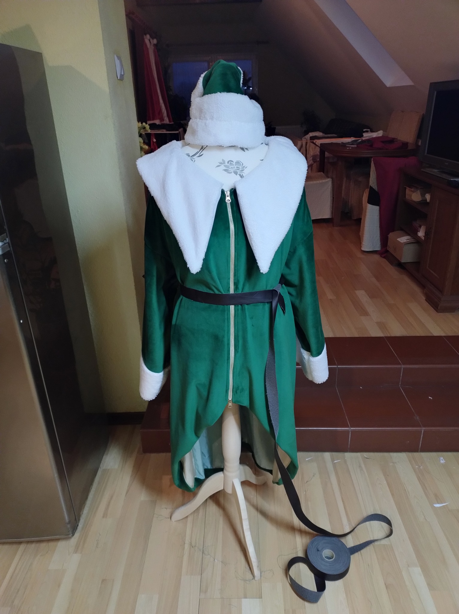 Niedokończony, zielony strój elfa z białym futerkiem na manekinie krawieckim, z czarnym paskiem leżącym na podłodze, w tle widoczny pokój na poddaszu.