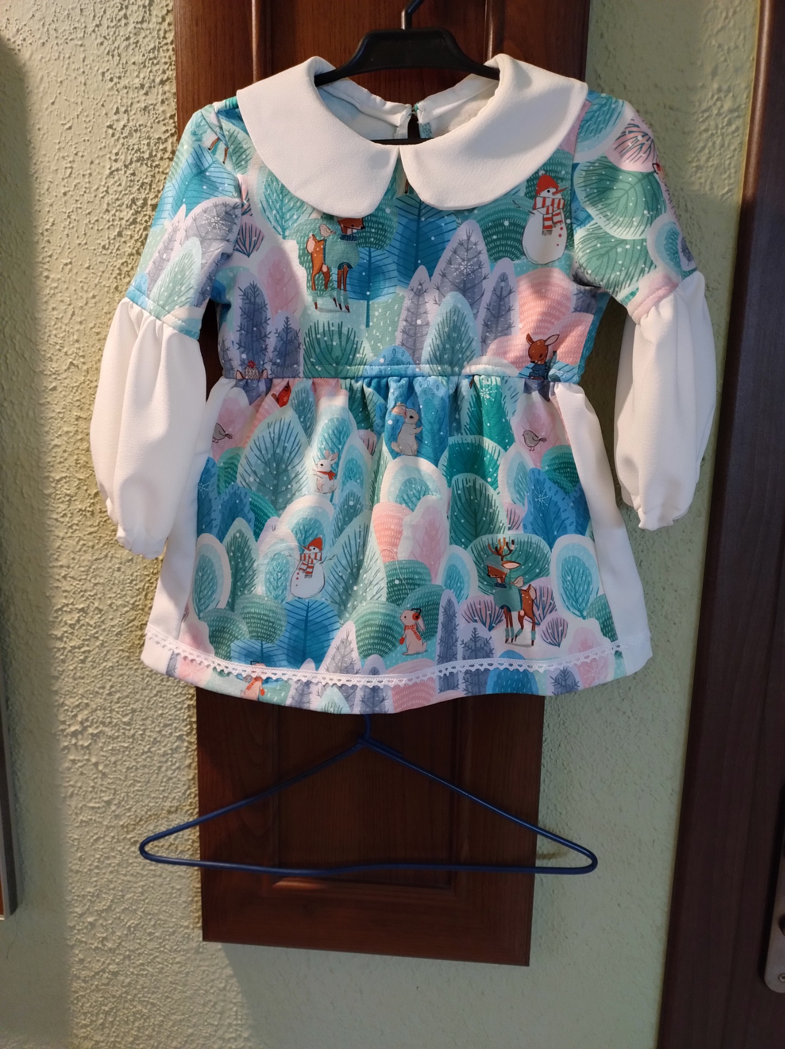 Sukienka dziecięca z motywem zimowego lasu i zwierząt, wykończona białym kołnierzykiem i koronką, wisząca na wieszaku.