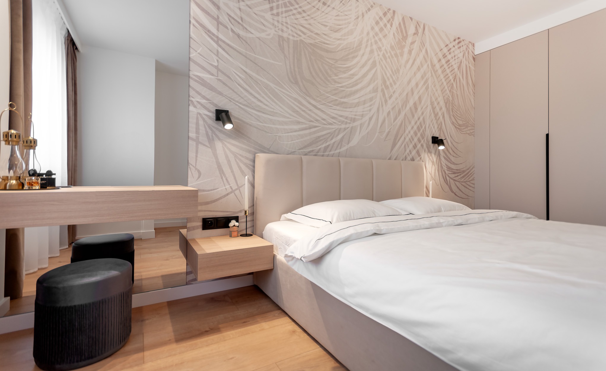 Nowoczesna sypialnia w neutralnych barwach z łóżkiem kontynentalnym, toaletką z lustrem, tapetą w motywy roślinne i drewnianą podłogą.