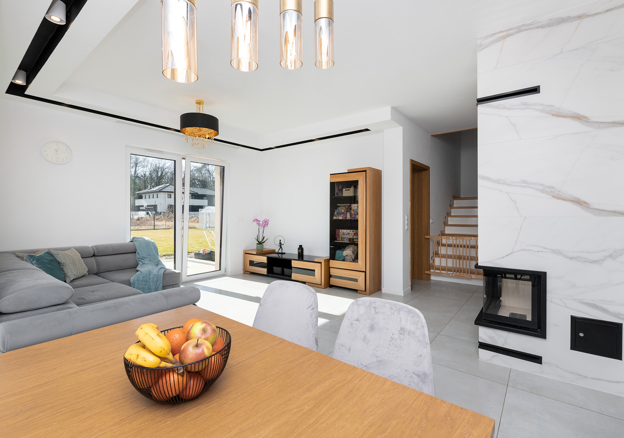 Jasne, przestronne wnętrze salonu z marmurowym kominkiem, drewnianym stołem z owocami w koszyku na pierwszym planie i szarą sofą w tle.