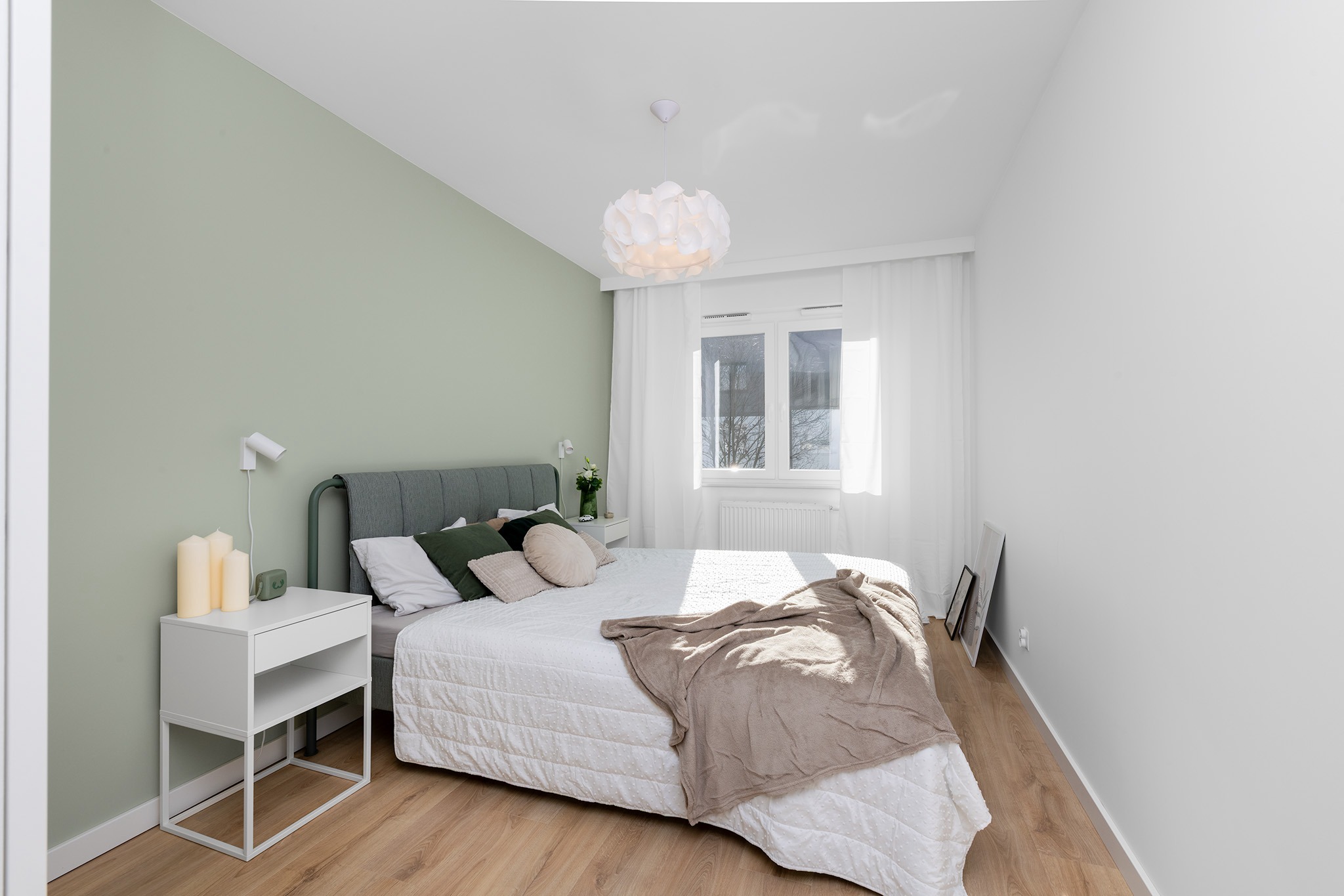Jasna sypialnia z łóżkiem z szarym zagłówkiem, białą pościelą, beżowym pledem i zielonymi poduszkami, białe szafki nocne z lampkami i świecami, drewniana podłoga, okno z białymi zasłonami, biała...