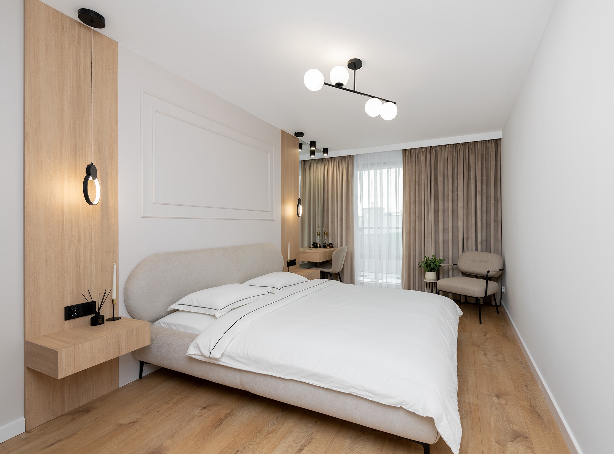 Jasna sypialnia z drewnianą podłogą, beżowym łóżkiem z białą pościelą, lampą wiszącą z drewnianym panelem, krzesłem obok okna z zasłonami i biurkiem w tle.