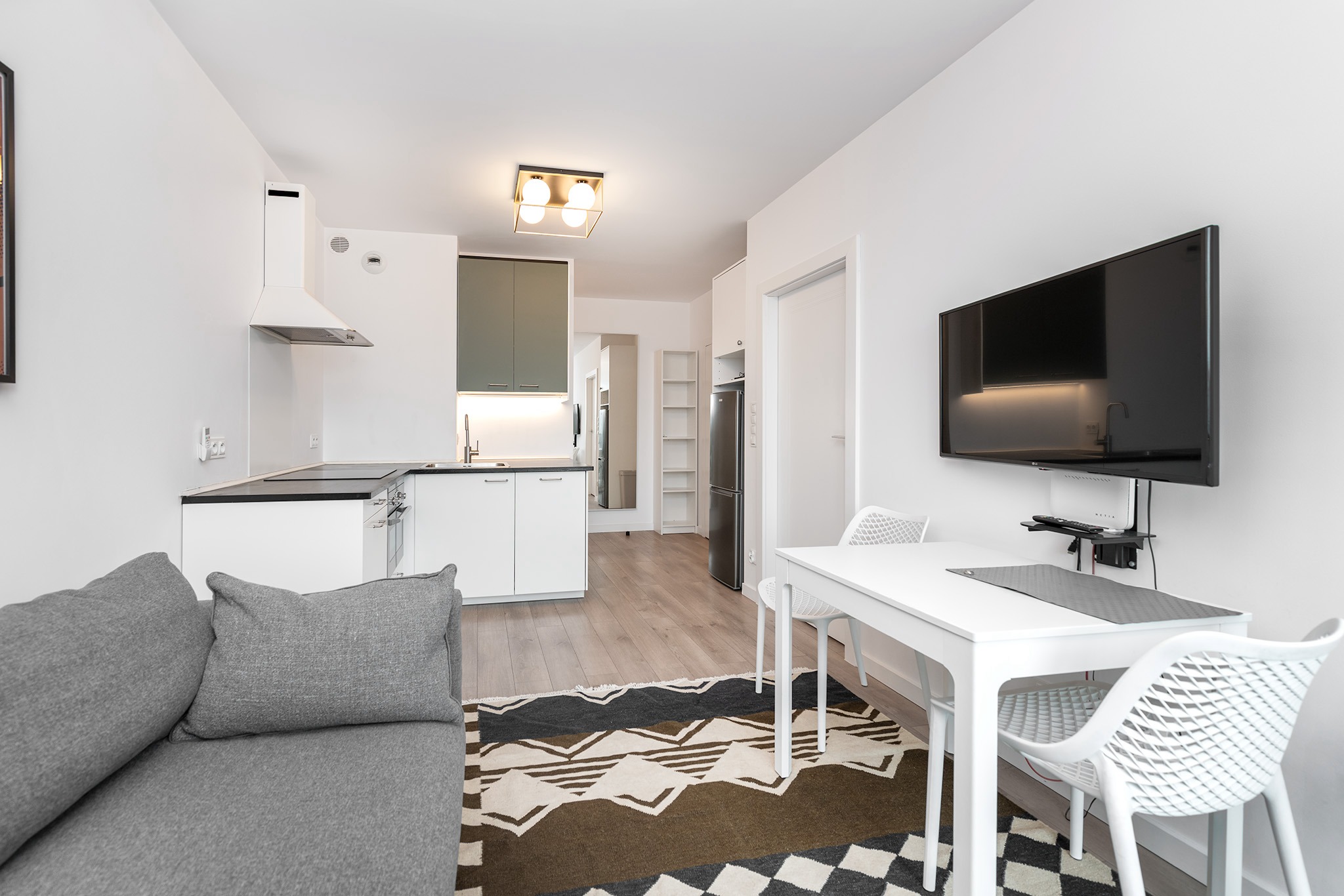 Wnętrze kawalerki z aneksem kuchennym, szarą sofą i białym stołem z krzesłami, oświetlone lampą sufitową z czterema kulami.