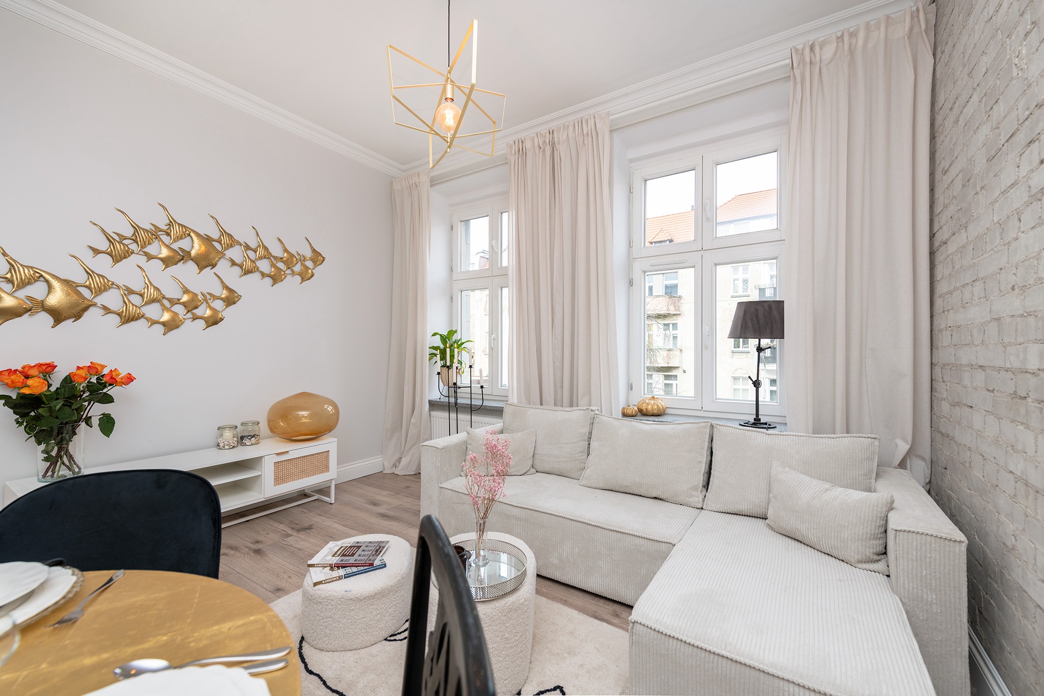 Jasne wnętrze salonu z narożną sofą z tkaniny typu sztruks, złotą dekoracją w kształcie ławicy ryb na ścianie i geometryczną lampą wiszącą.