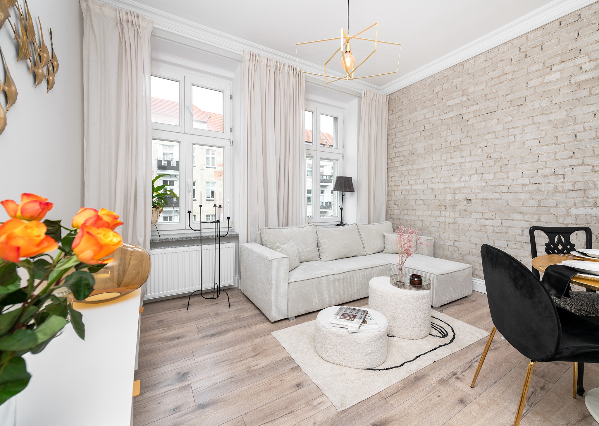 Jasny salon z sofą z tkaniny w prążki, dwoma okrągłymi pufami na dywanie z czarnym wzorem, ścianą z białej cegły i geometryczną lampą wiszącą.