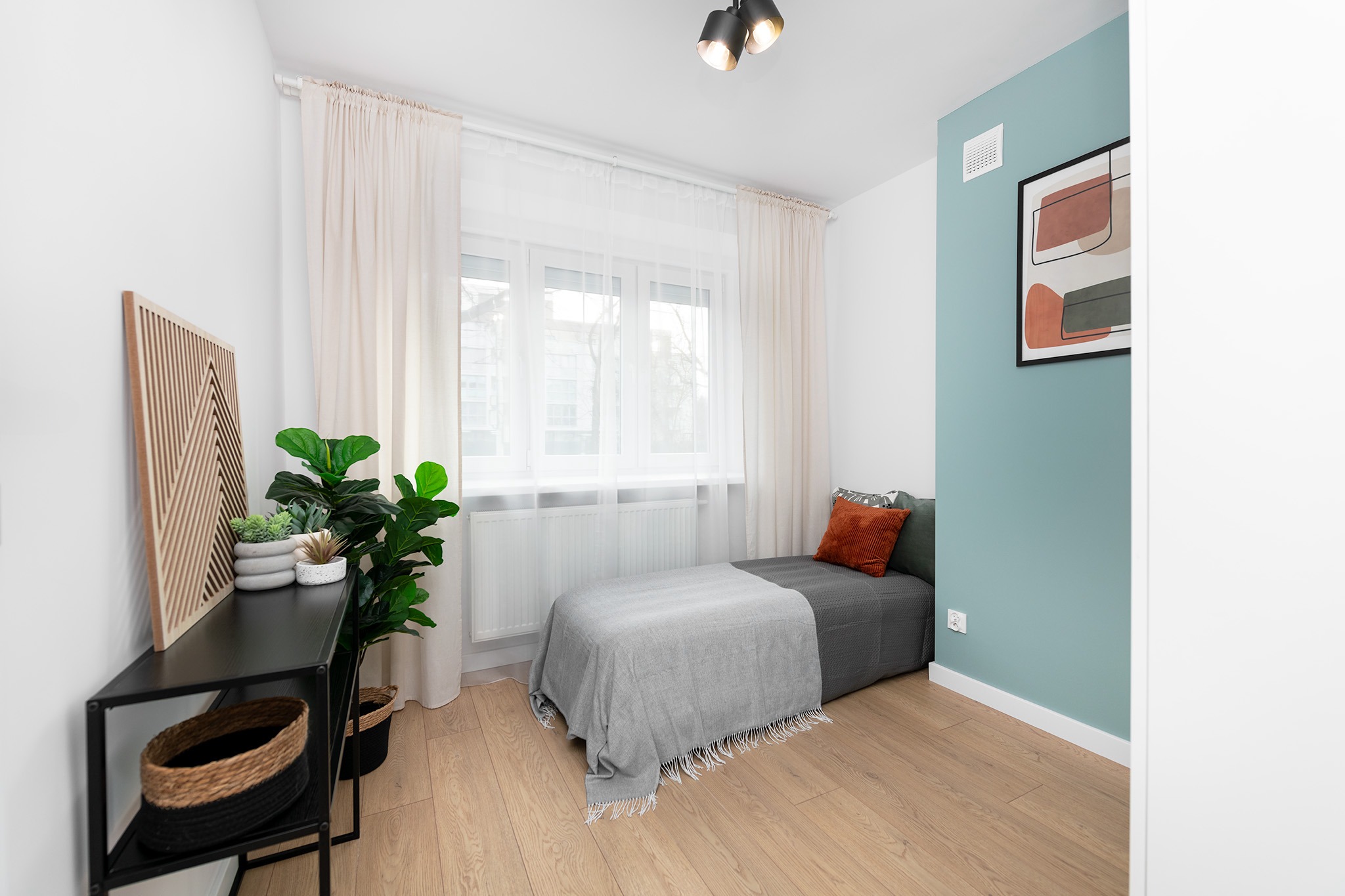 Pokój młodzieżowy z pojedynczym łóżkiem, turkusową ścianą z abstrakcyjnym obrazem, czarną konsolą z drewnianą dekoracją i roślinami doniczkowymi.