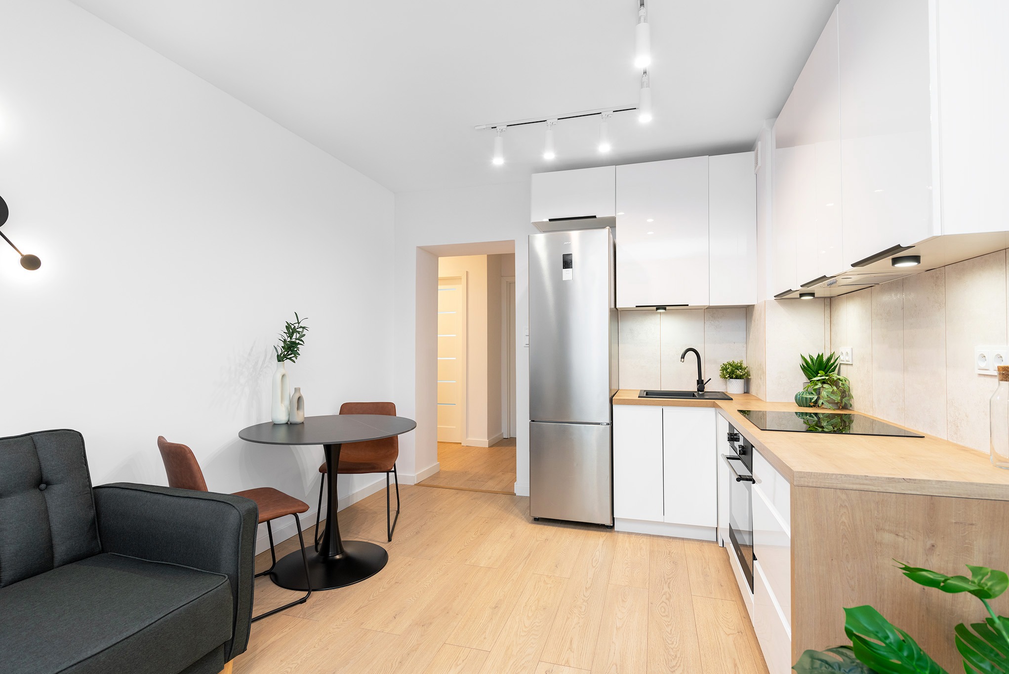 Nowoczesne wnętrze mieszkania: biała kuchnia z drewnianym blatem, srebrna lodówka, czarny stół z brązowymi krzesłami i szara sofa, oświetlenie punktowe.